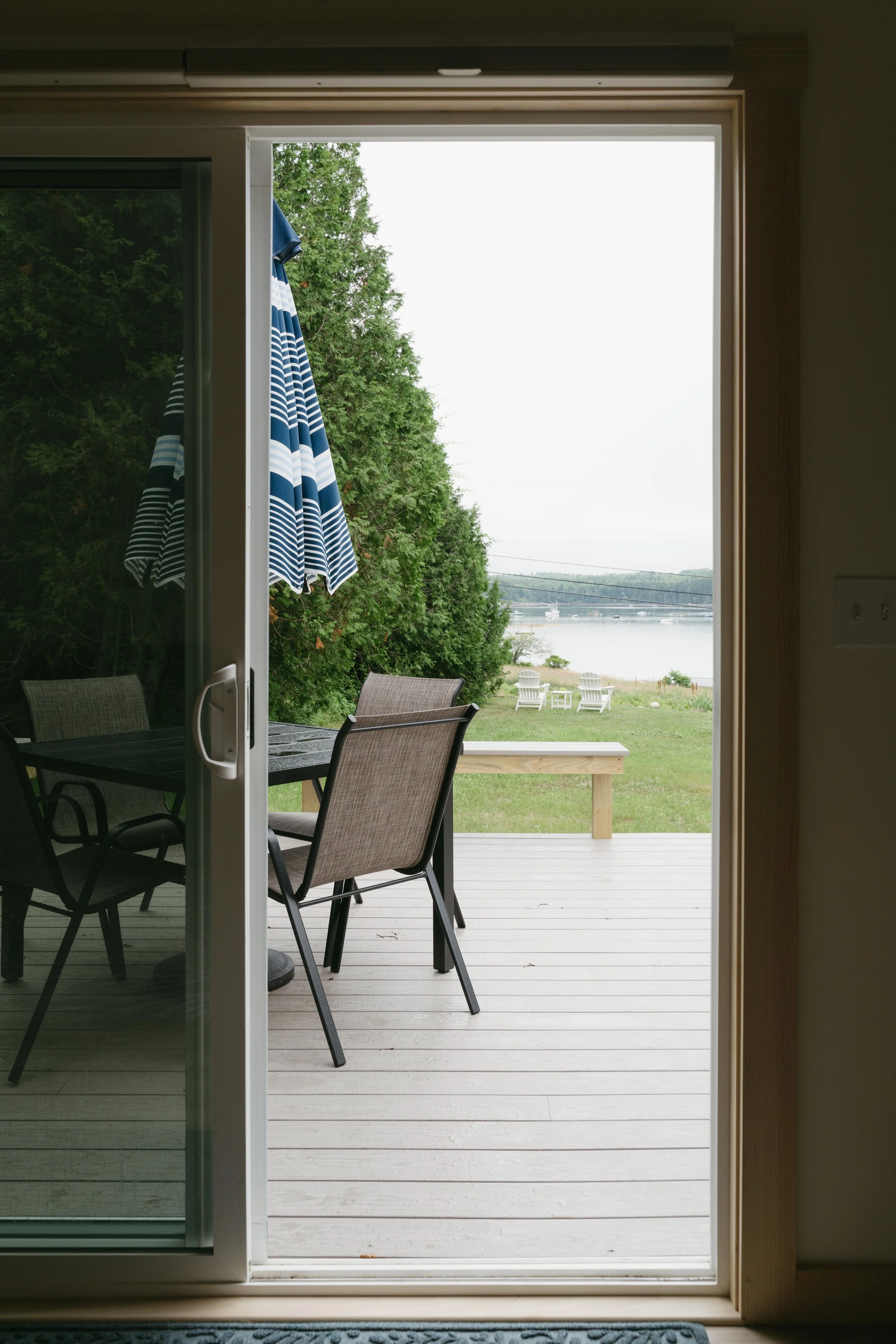 View through a sliding glass door onto a wooden deck with outdoor chairs, trees, and a body of water in the distance.