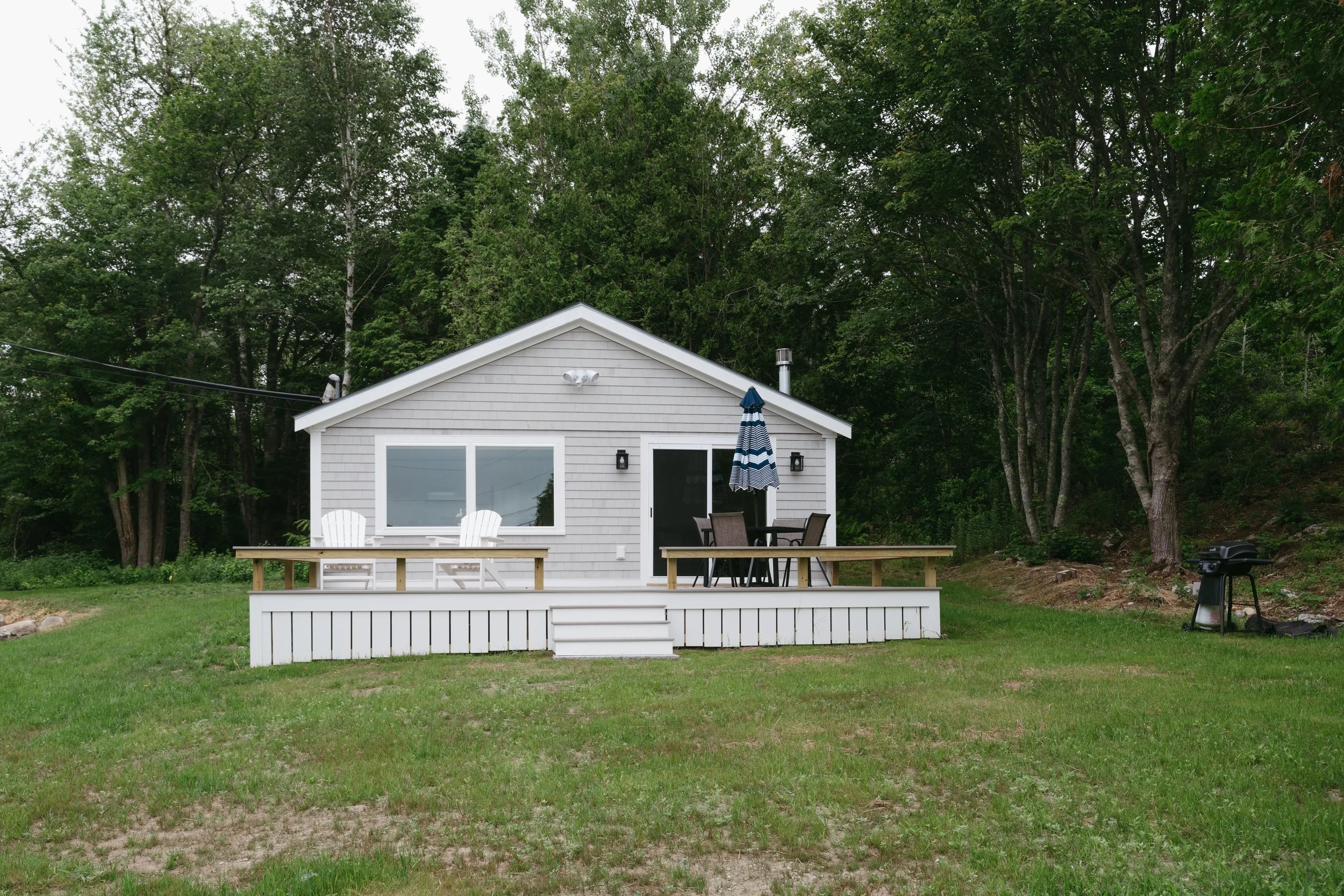 A small gray house with a wooden deck, two white Adirondack chairs, a black patio table with chairs, and a blue and white striped umbrella, surrounded by green trees and grass, with a barbecue grill on the right side.