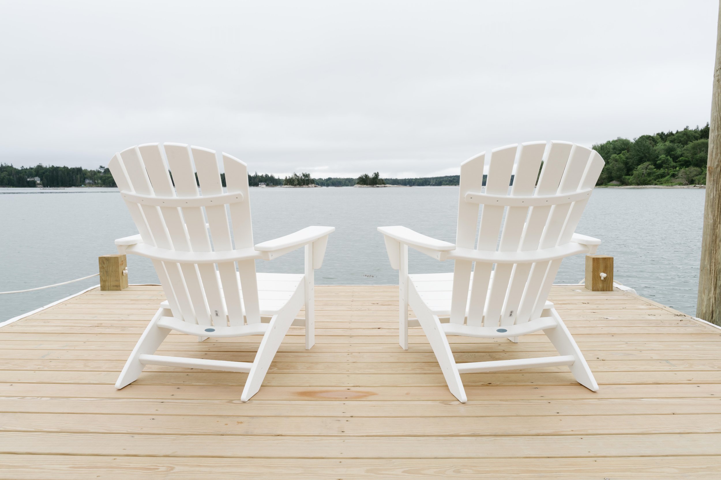 Two white Adirondack chairs on a wooden dock facing a body of water with distant trees and an overcast sky.