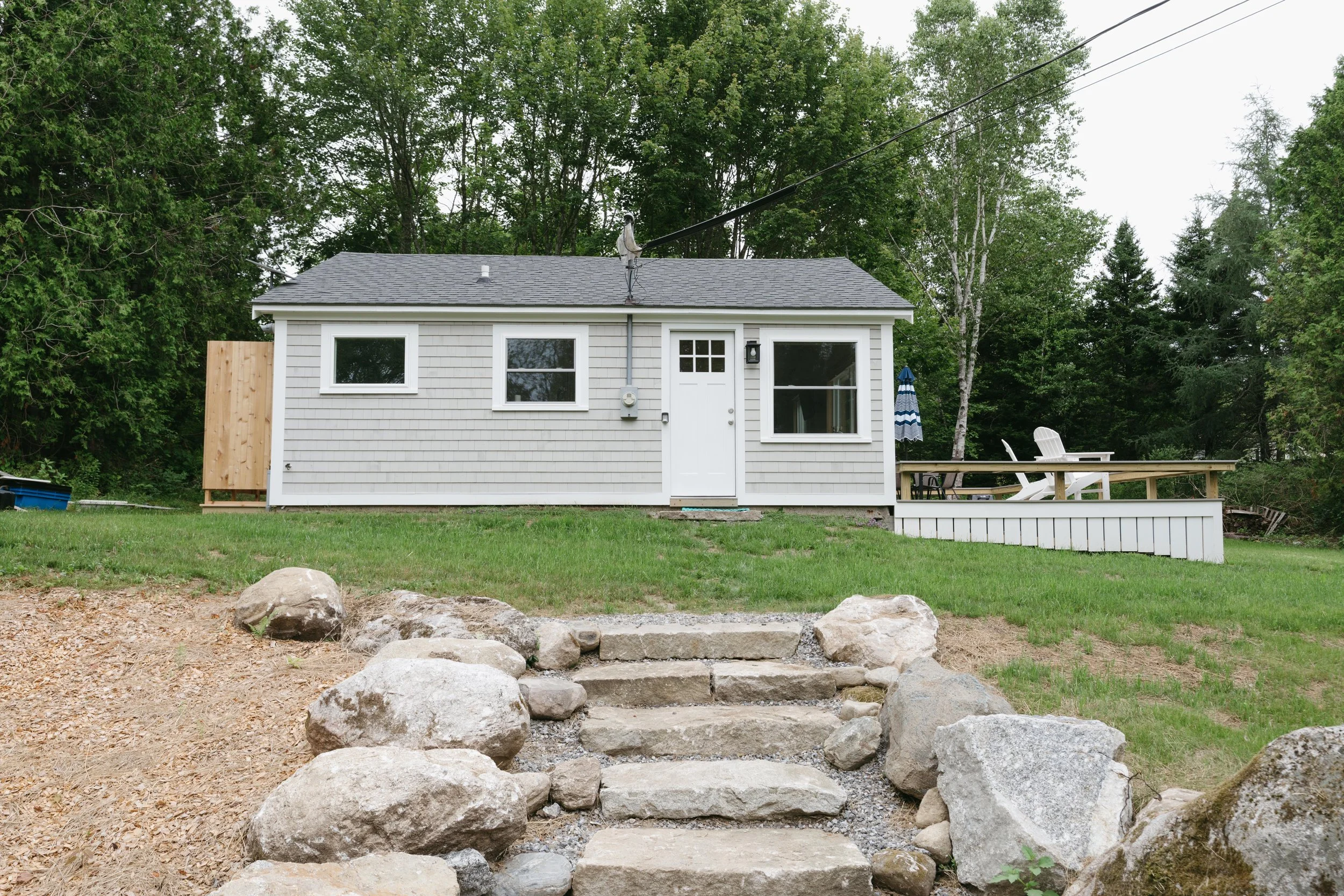 A small, white, single-story house with a gray roof, a door with a small window, and three windows. There is a deck with white chairs and a blue and white umbrella. The yard has a stone staircase leading up to the house, large rocks on either side, a