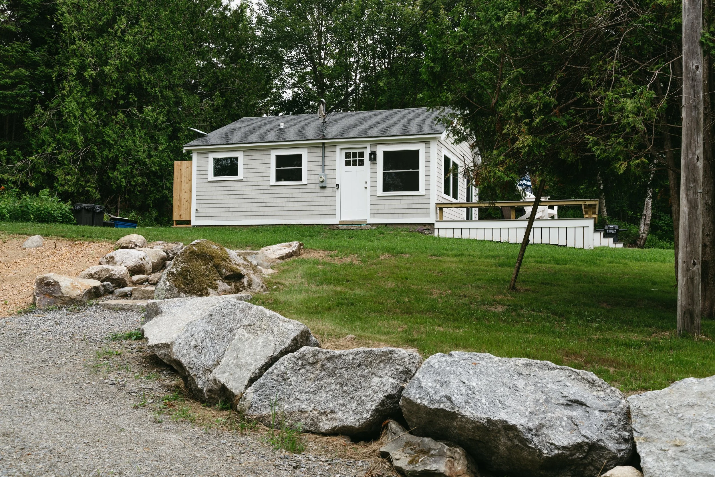 A small gray house with white trim, a black roof, and a front door, situated on a grassy hill with large rocks in the foreground and surrounded by trees.