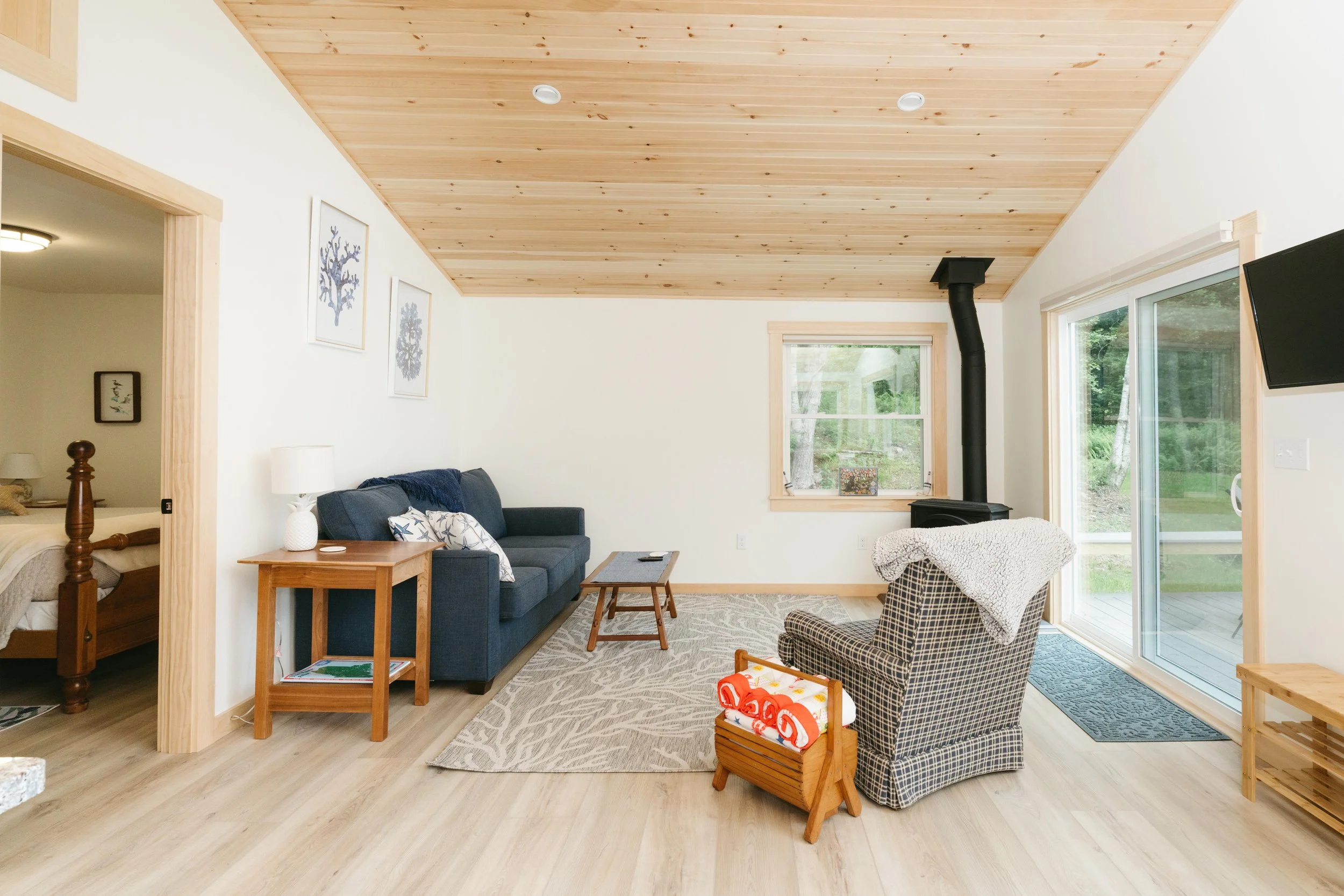 Living room with a blue sofa, checkered armchair, wooden coffee table, tile rug, wood ceiling, sliding glass door, window, and a television mounted on the wall.