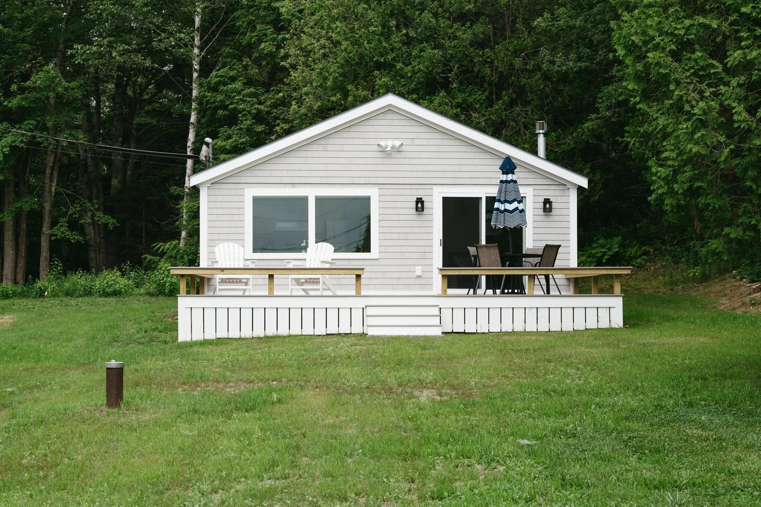 A small white house with a deck, two Adirondack chairs, a dining table with chairs and a blue umbrella, set against a background of dense green trees.
