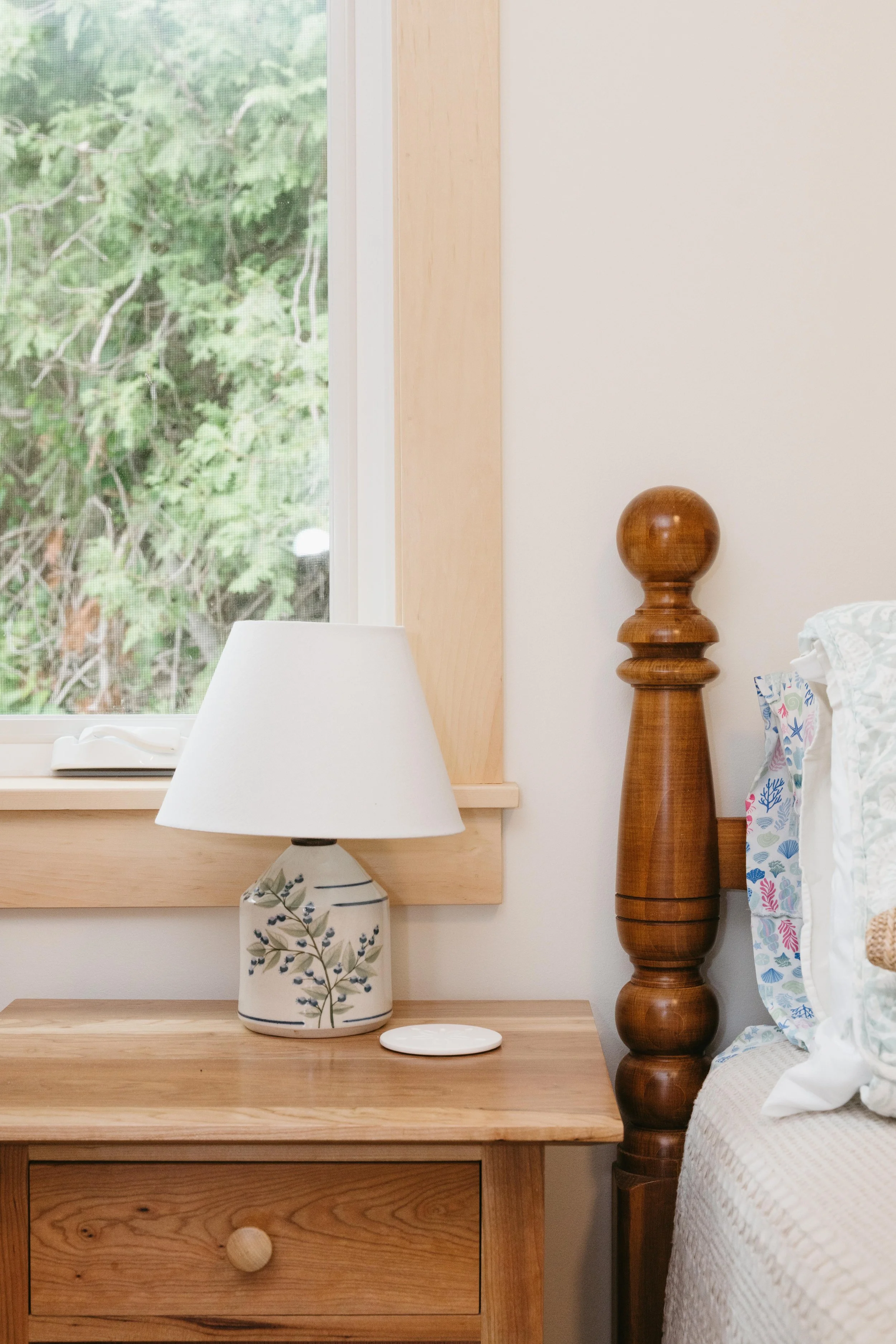 Close-up of a nightstand with a ceramic table lamp, a window with a view of green trees, and part of a wooden bedpost with a decorative turn.