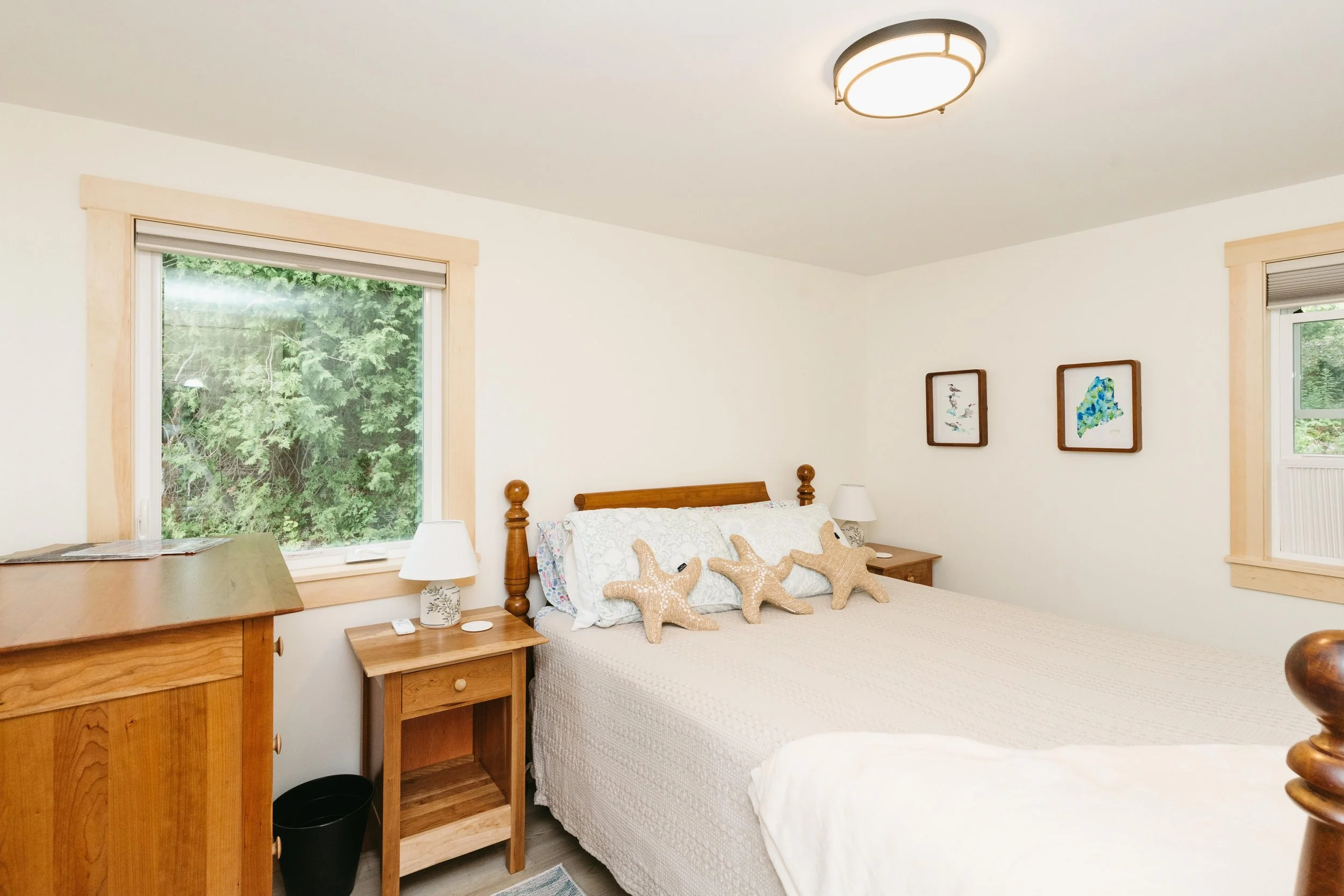 A bedroom with a wooden bedframe and starfish pillows, two windows with green foliage outside, two small side tables with lamps, and two framed pictures on the wall.