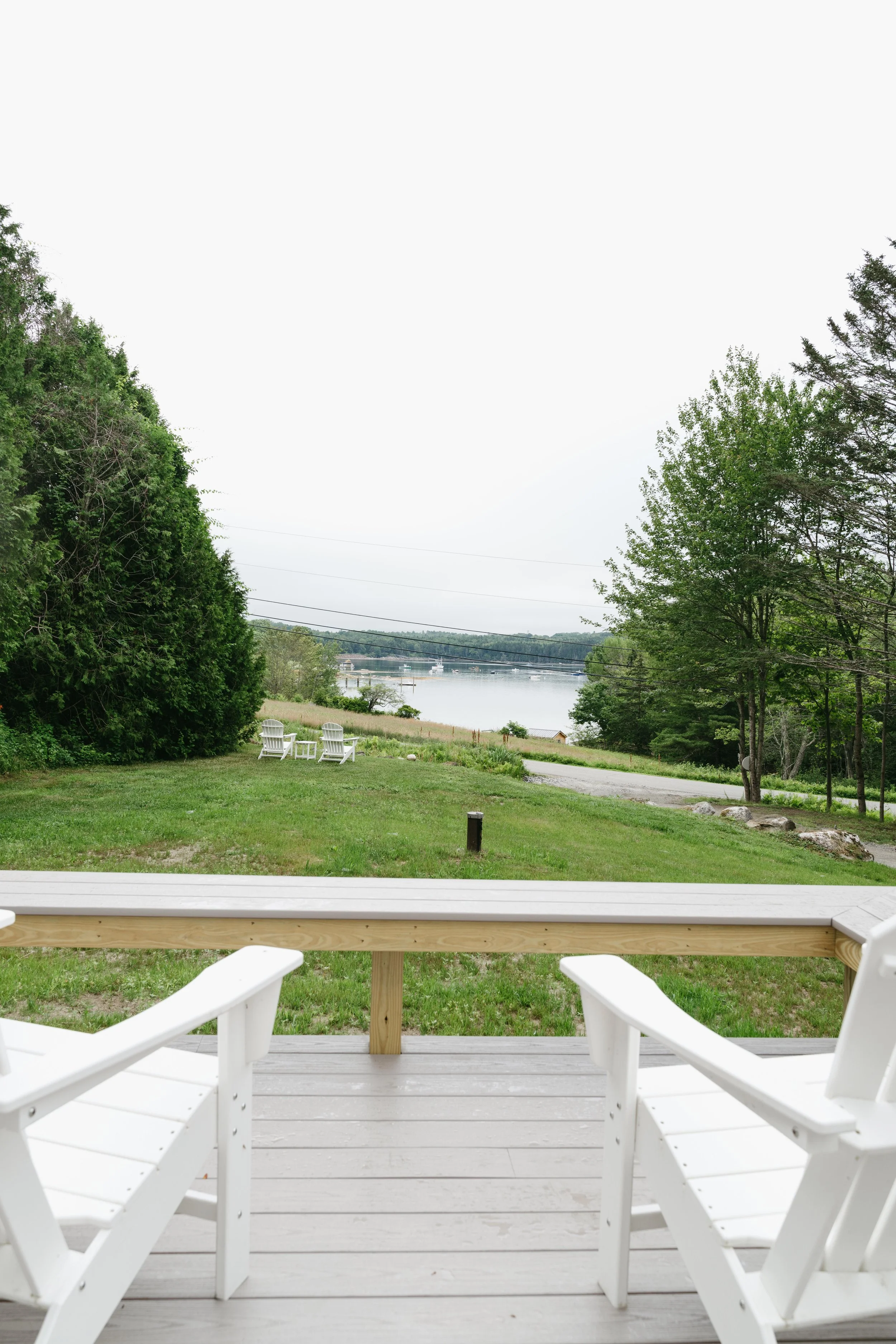 View from a porch overlooking a green yard with two white chairs, trees, a body of water, and a bridge in the distance on a cloudy day.