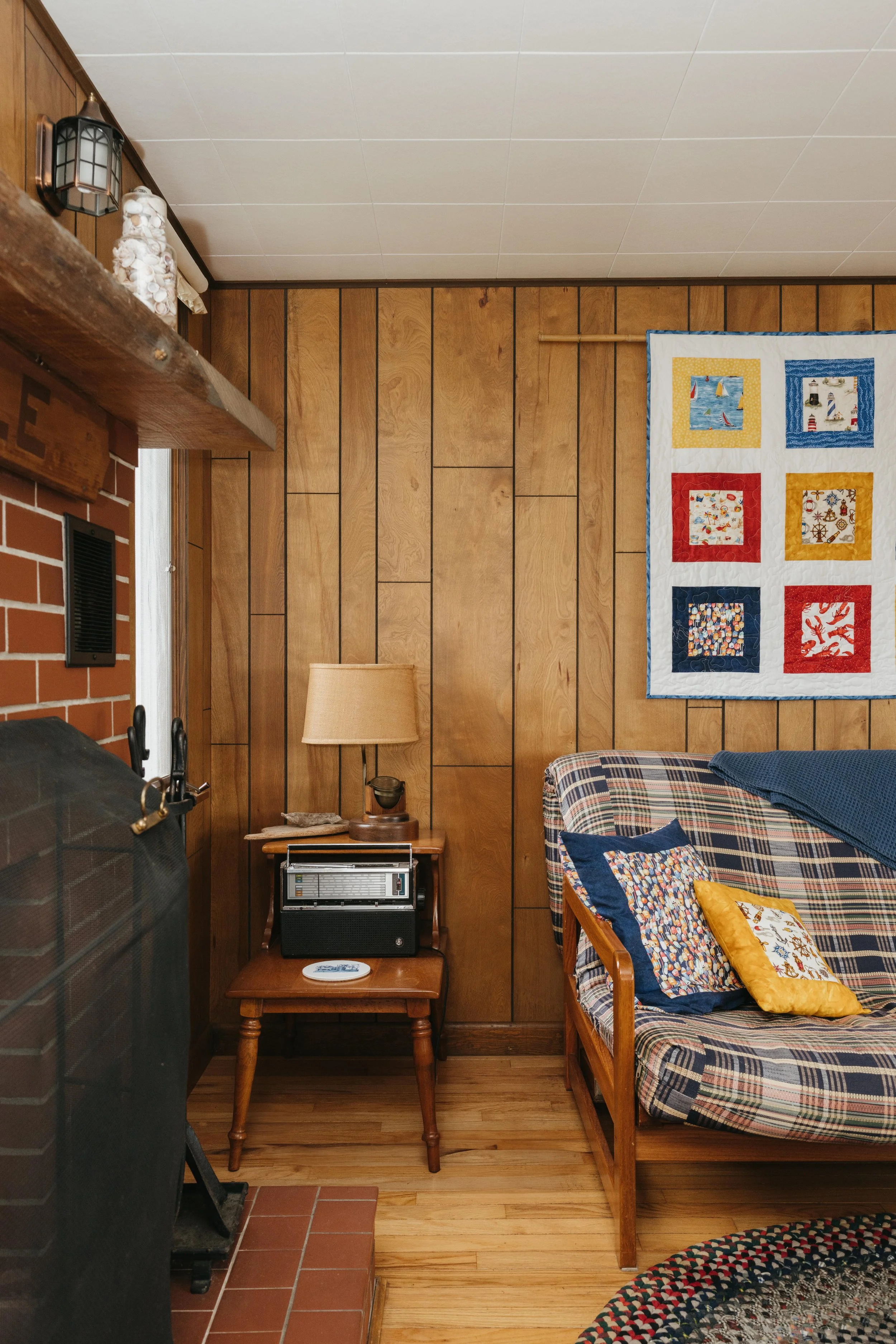 Living room corner with wood-paneled wall, colorful quilt hanging, plaid sofa with decorative pillows, wooden side table with lamp and radio, brick fireplace, and corner of a rug.