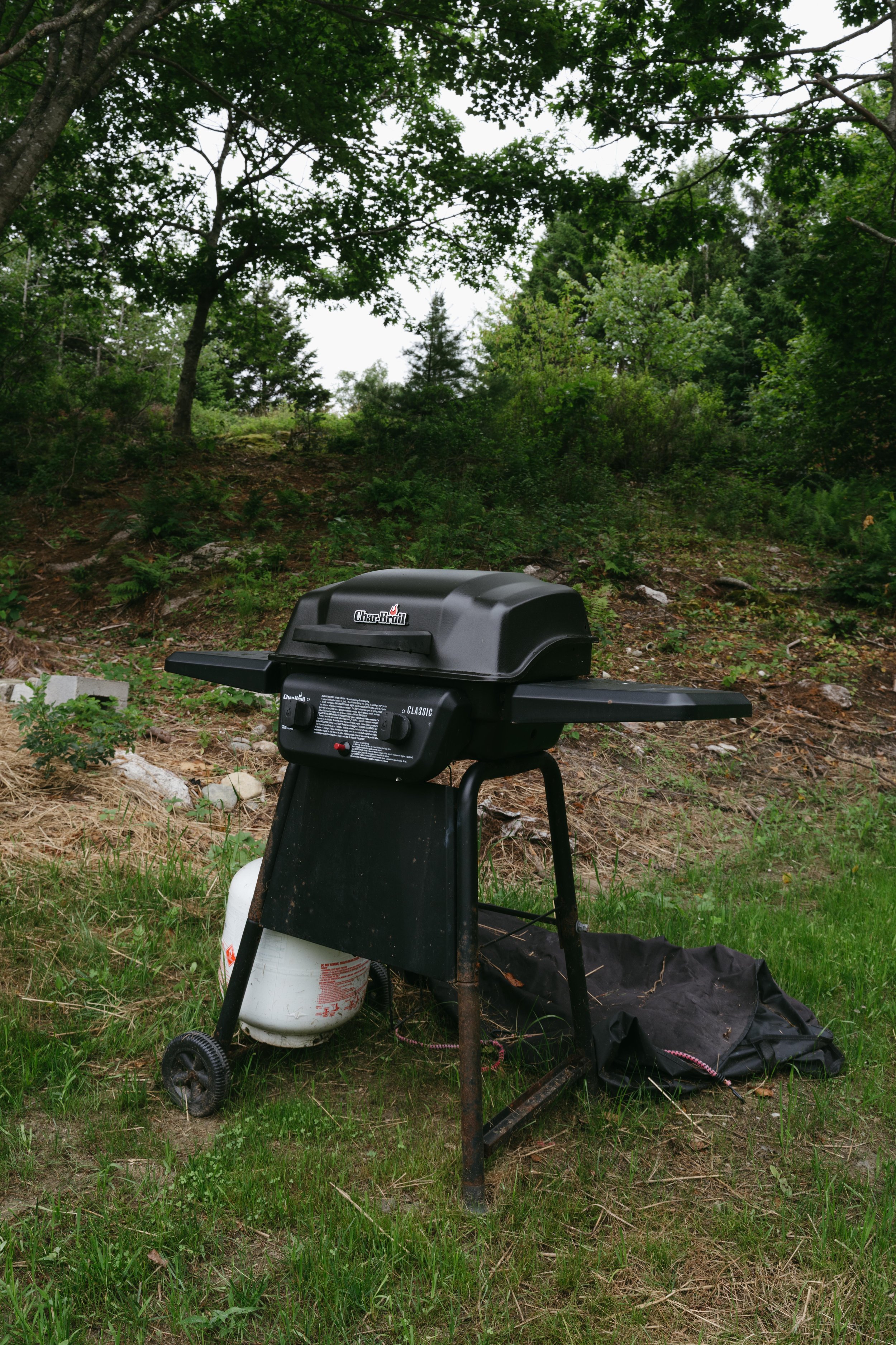 A black Char-Broil gas grill on a stand with a propane tank underneath, set on a grassy area outdoors surrounded by trees and bushes.