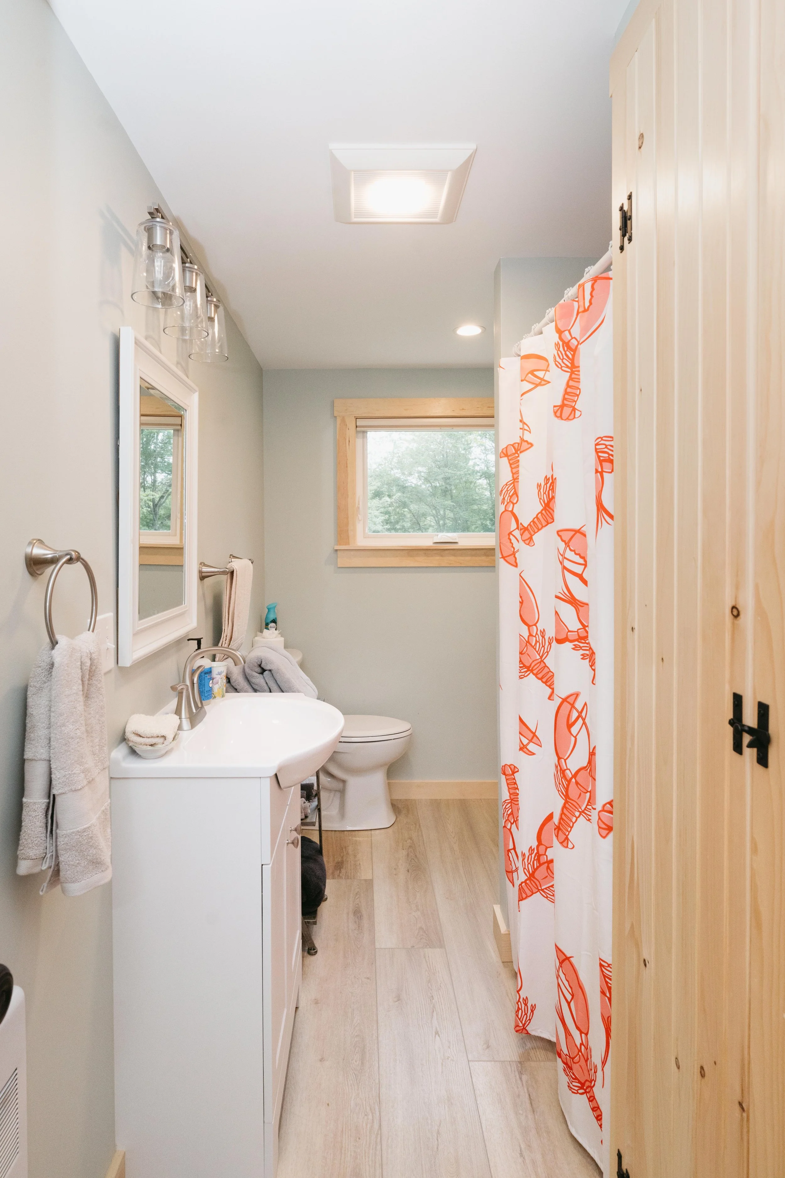 A bathroom with light-colored walls, a small window, a white sink with a mirror, a toilet, wooden flooring, wooden trim around the window, and a shower curtain with orange lobsters on it.