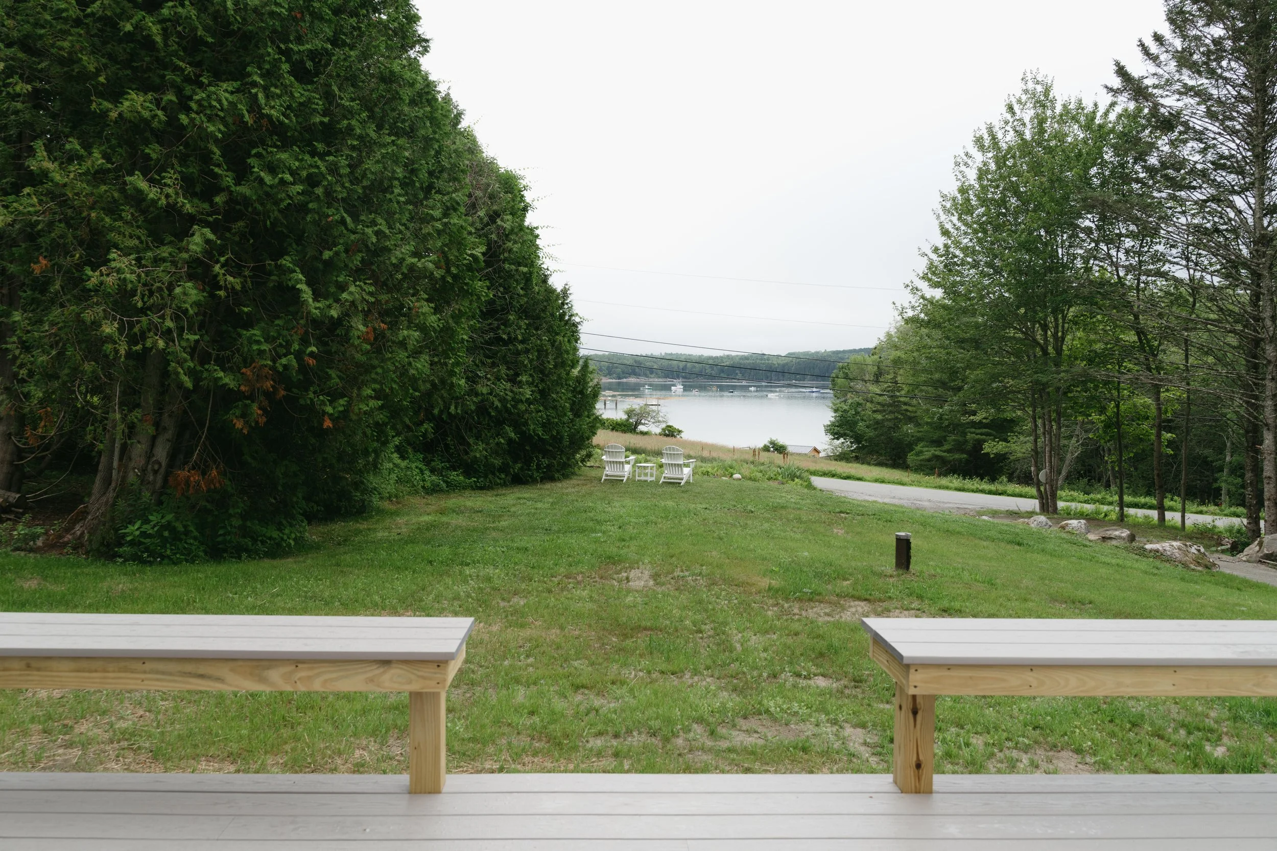 View of a grassy yard with two white Adirondack chairs facing a lake, surrounded by trees on a cloudy day.