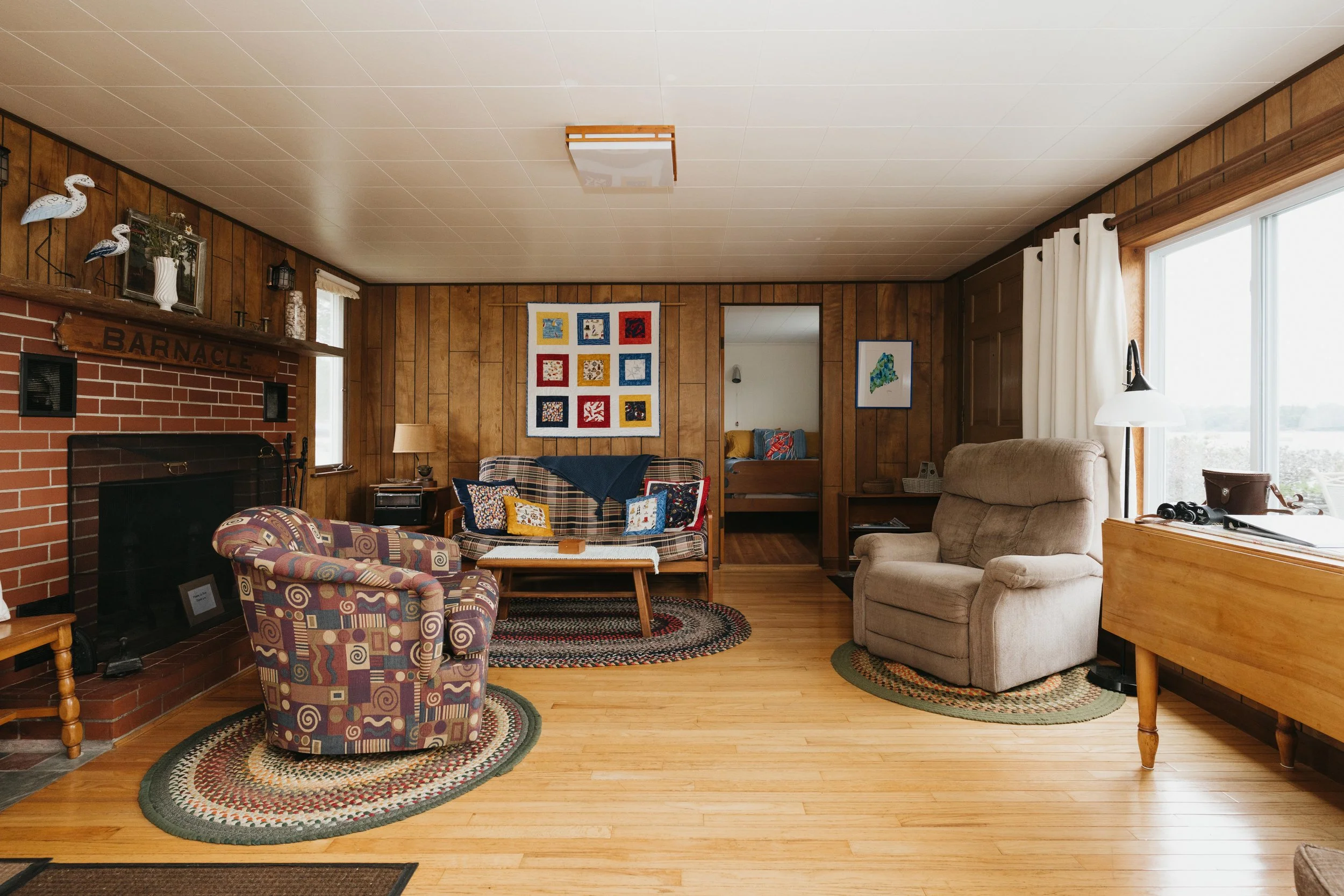 Cozy living room with wood-paneled walls, a brick fireplace with a sign reading 'Barnacle,' colorful patchwork quilted wall art, armchairs, a beige recliner, a sofa with patterned cushions, a wooden coffee table, and large windows with white curtains