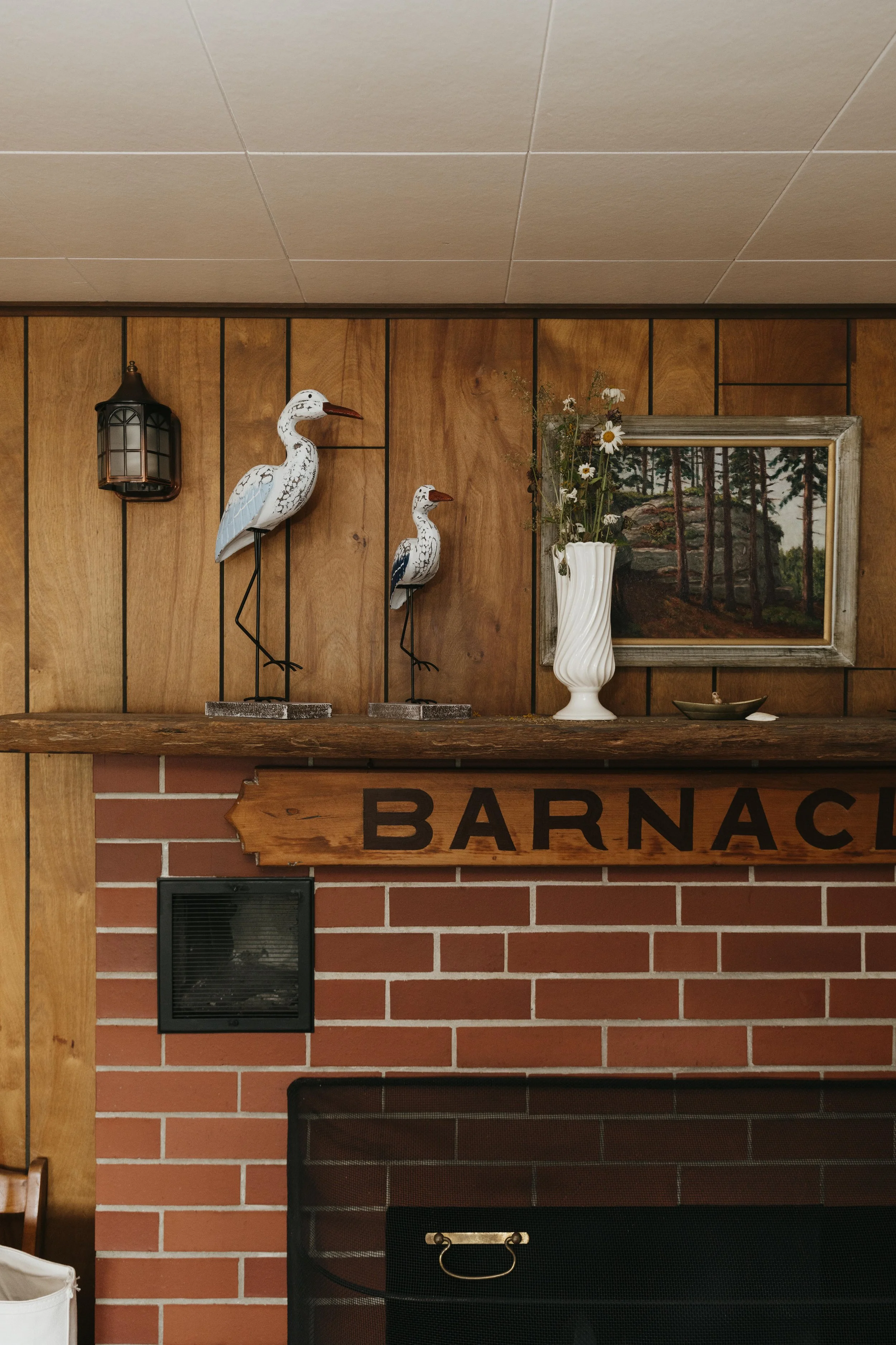 Decorative wall with wooden paneling and brick fireplace, featuring two bird sculptures on a wooden shelf, a white vase with flowers, a landscape painting, and a wall-mounted lantern.
