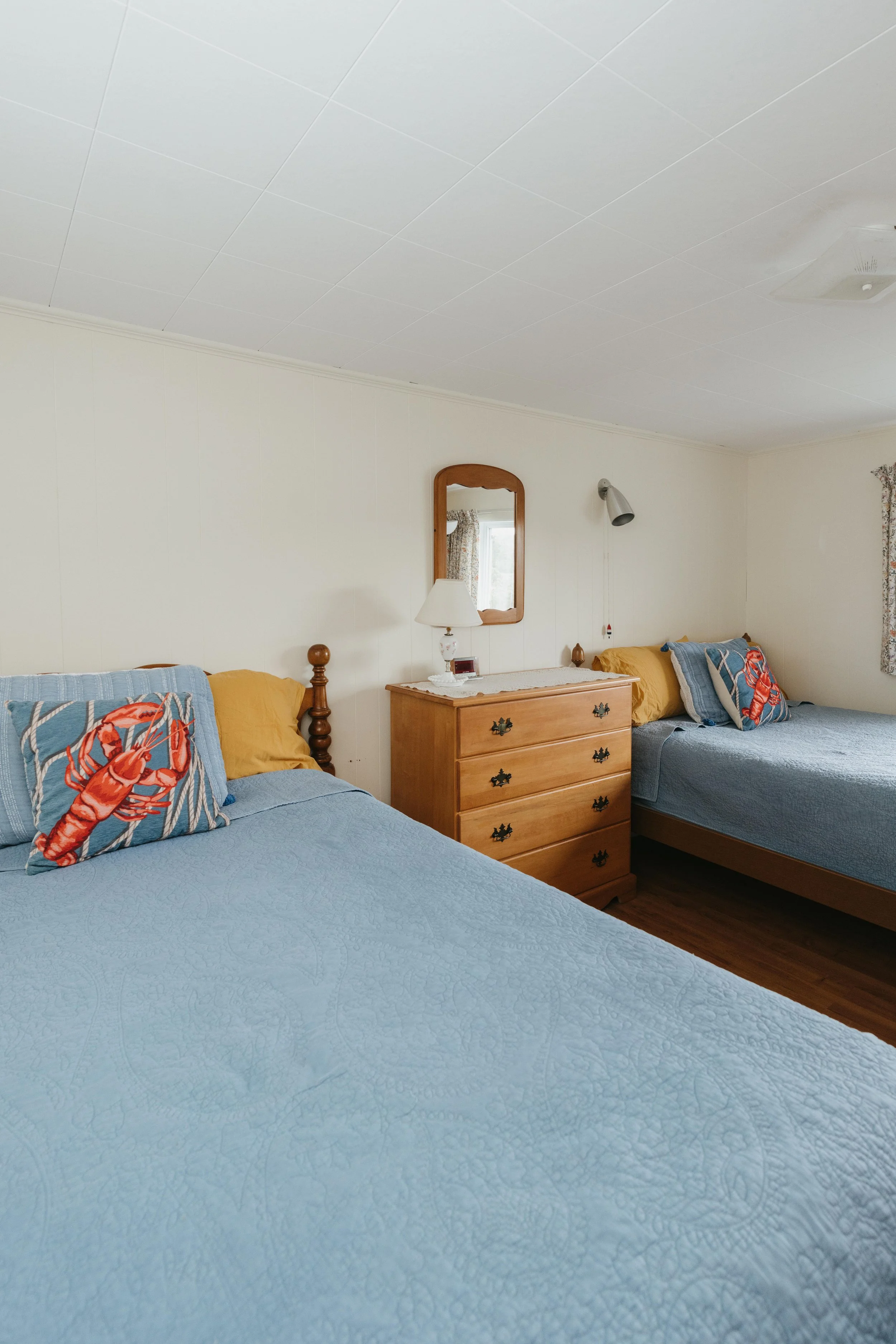 A bedroom with two twin beds covered in light blue bedspreads, lobster-themed pillows, a wooden dresser between the beds, a mirror above the dresser, a table lamp, and wall-mounted reading lights.