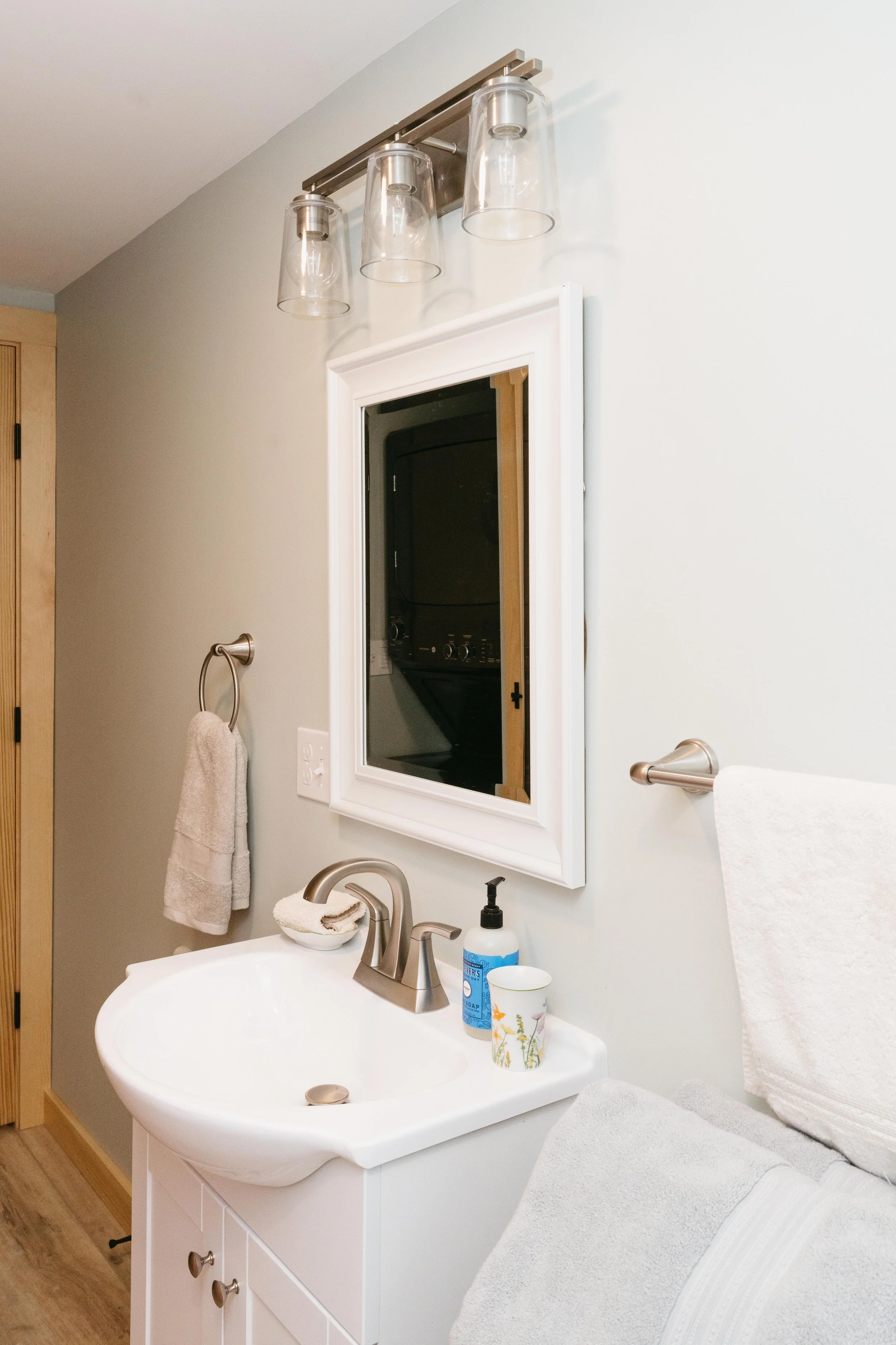 Bathroom vanity with a small sink, mirror, wall-mounted light fixture, soap dispenser, a paper cup, and towel hooks with towels.