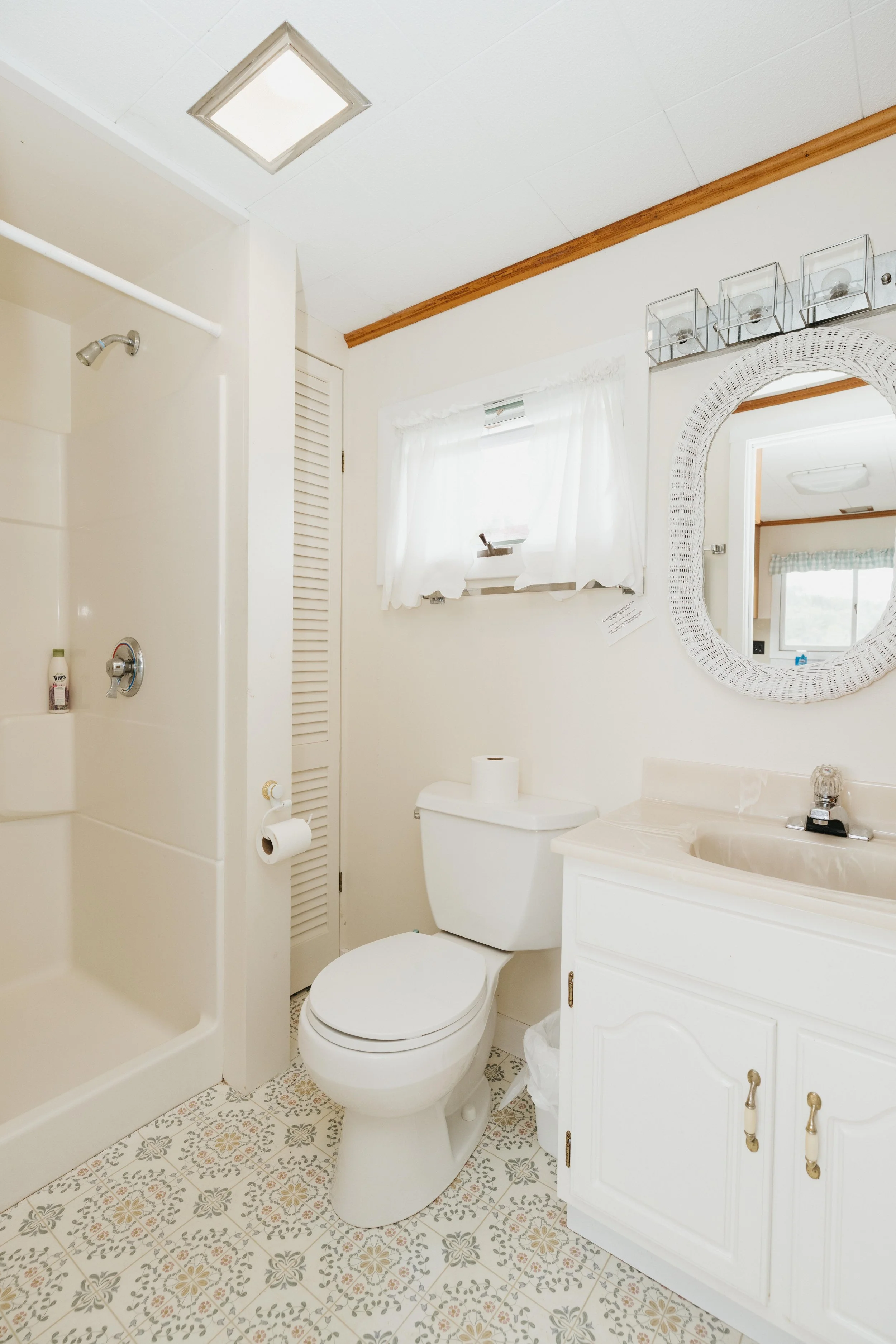 A small bathroom with a shower on the left, a toilet in the center, and a white vanity with a sink and oval mirror on the right. There is a small window with white curtains above the toilet and a decorative shelf with glass containers on the wall.
