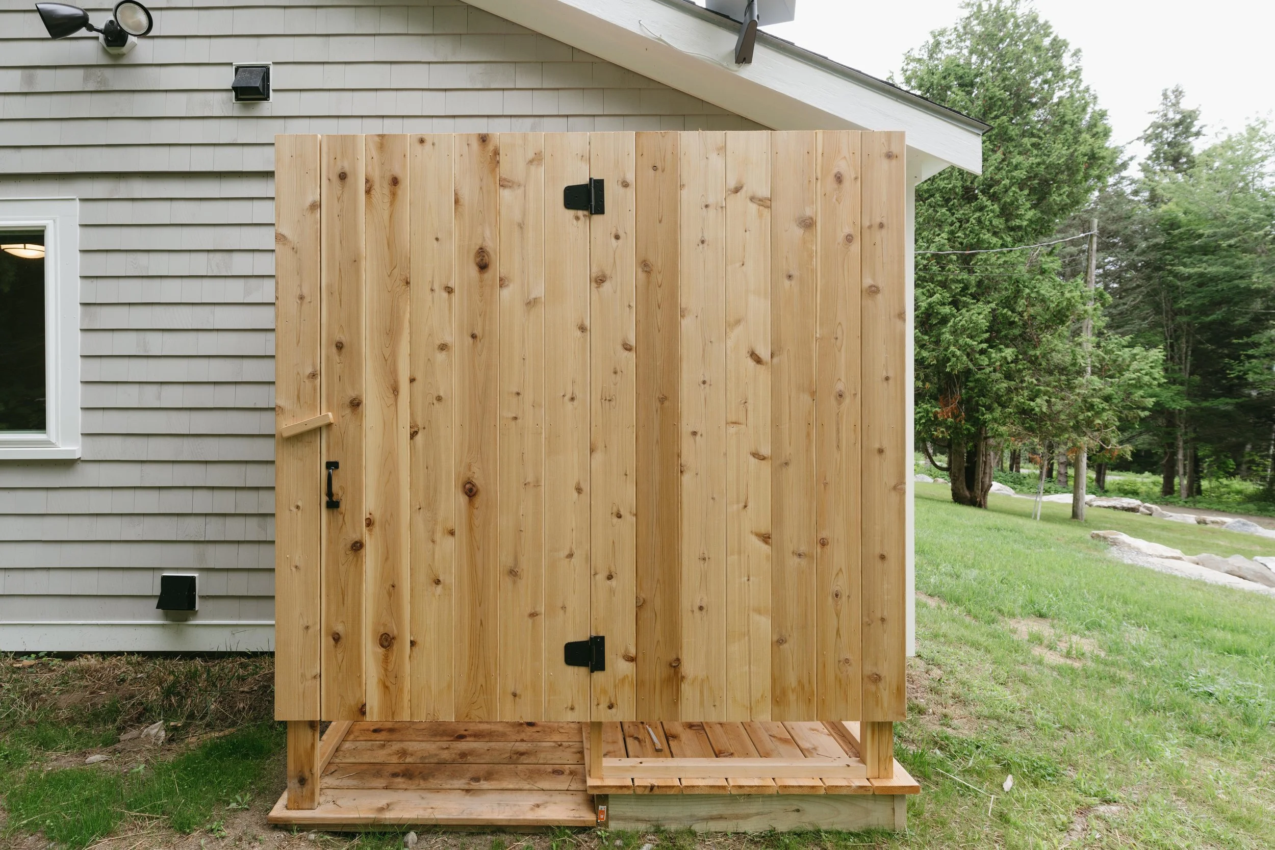 A wooden outdoor shower enclosure with a door and a built-in bench on a small wooden platform next to a house with gray horizontal siding, in a grassy yard with trees in the background.
