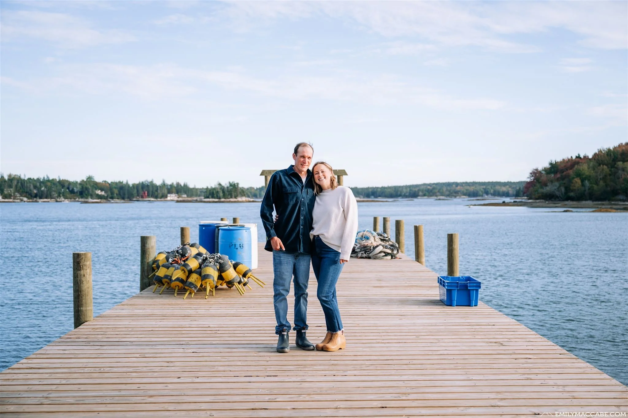 A couple standing on a wooden dock by a body of water, with equipment such as buoys, barrels, and a blue crate around them, and trees in the background.