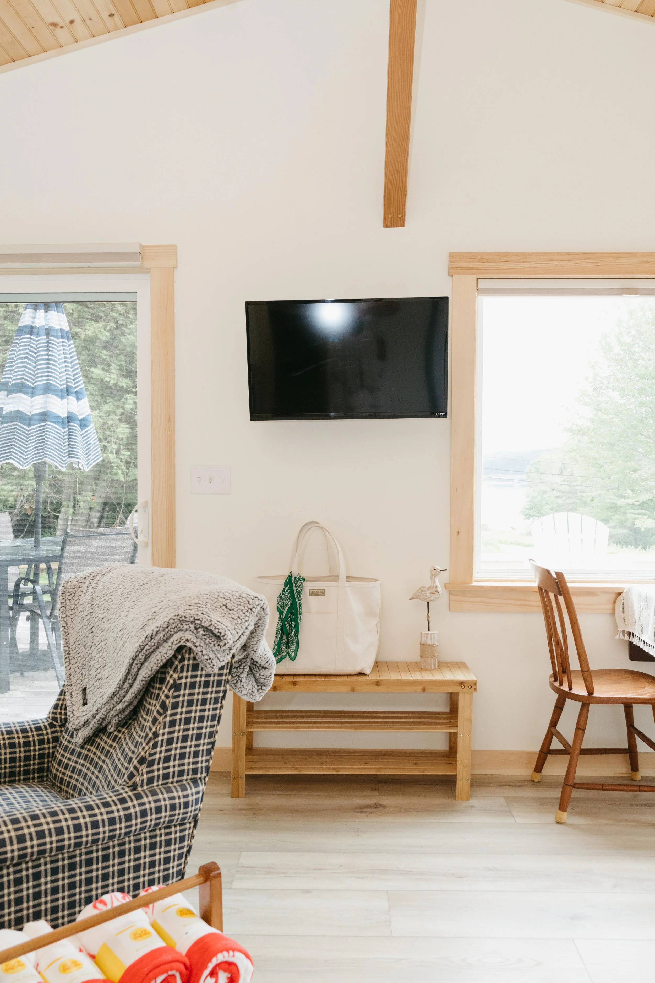 A living room corner with a wall-mounted flat-screen TV above a wooden bench, a window with light wood trim, a small white bag on the bench, a bird figurine, and a wooden chair next to the window.