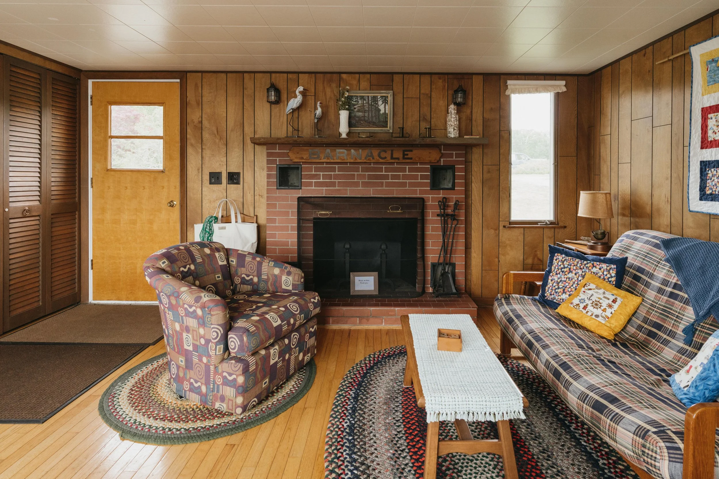 Living room with wood-paneled walls, a brick fireplace with a wooden mantel, and various decorations including a sign that says 'BARNACLE'; patterned armchair, checkered sofa with pillows, small wooden coffee table with a white runner, side table wit