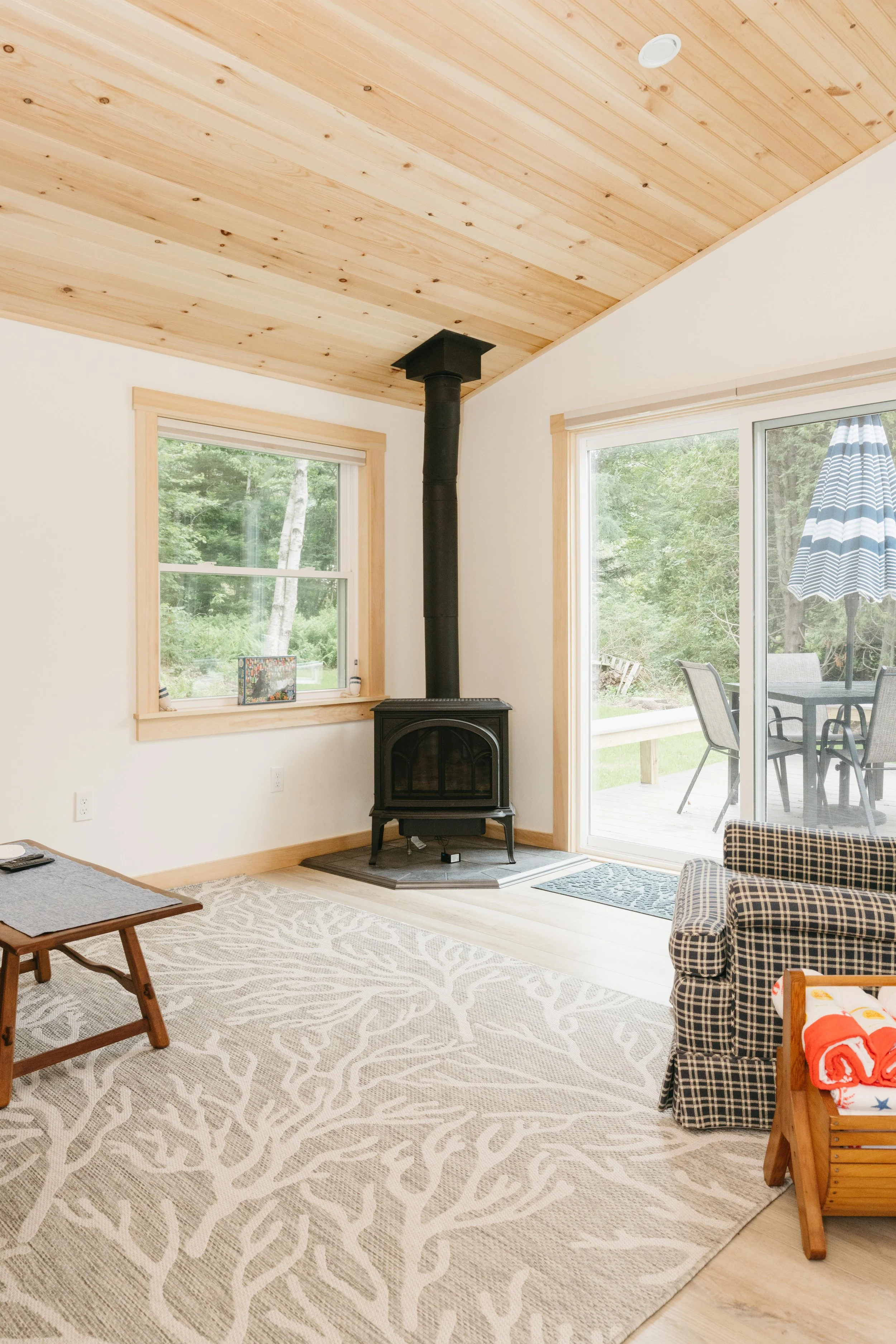 Living room with wood ceiling, black stove, sliding glass door leading to patio, window showing trees, armchair with checkered fabric, rug with coral design, and patio furniture outside.