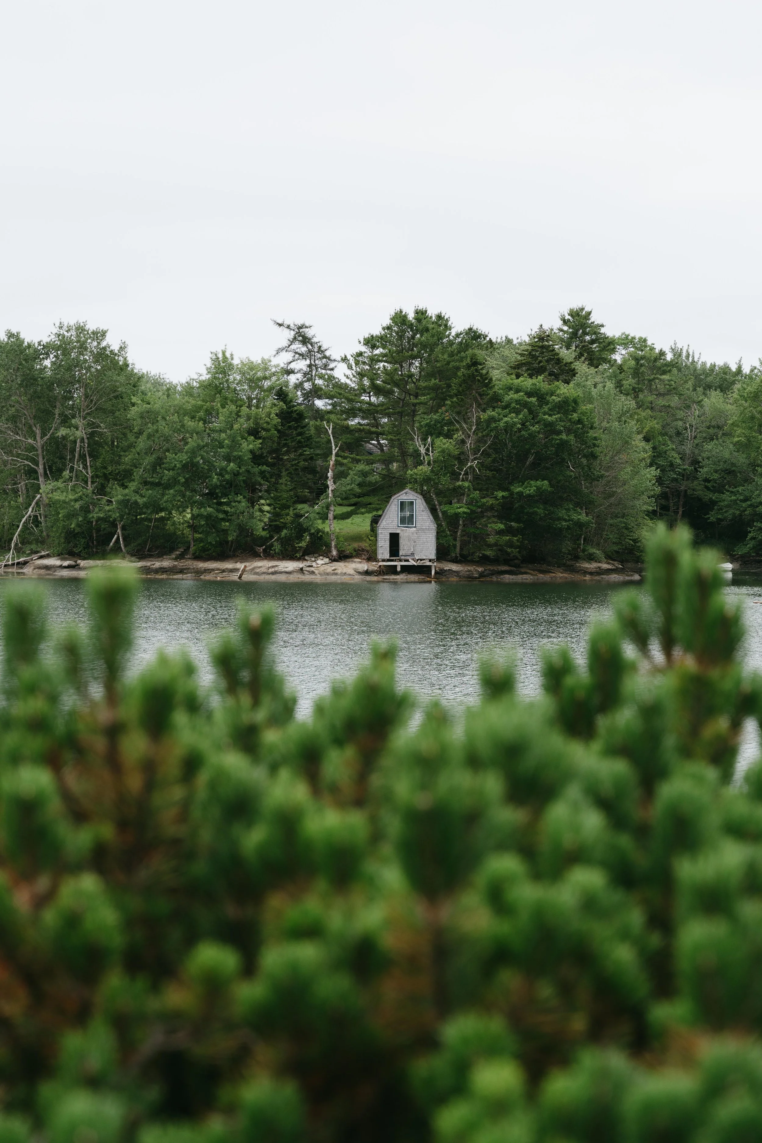 A peaceful lakeside scene with a small gray boathouse on the shore, surrounded by green trees, with water in the foreground and an overcast sky.