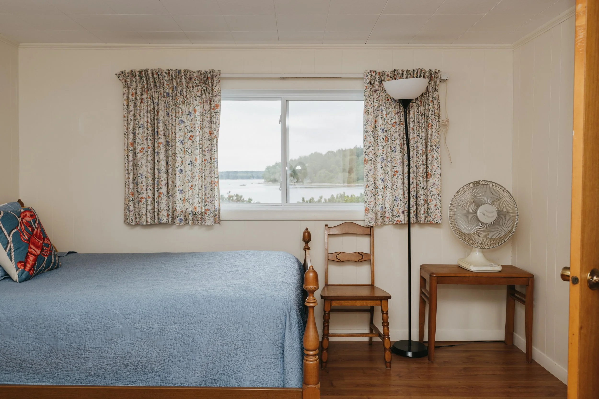A bedroom with a bed, a window with floral curtains, a wooden chair, a small wooden table with a fan, a floor lamp, and wooden flooring.