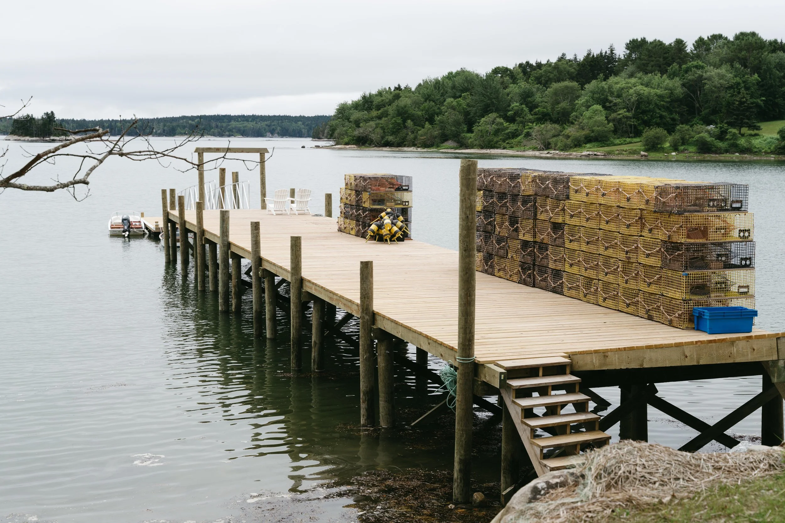 A wooden dock extends over a body of water with stacks of lobster traps on it, surrounded by green trees and overcast sky.