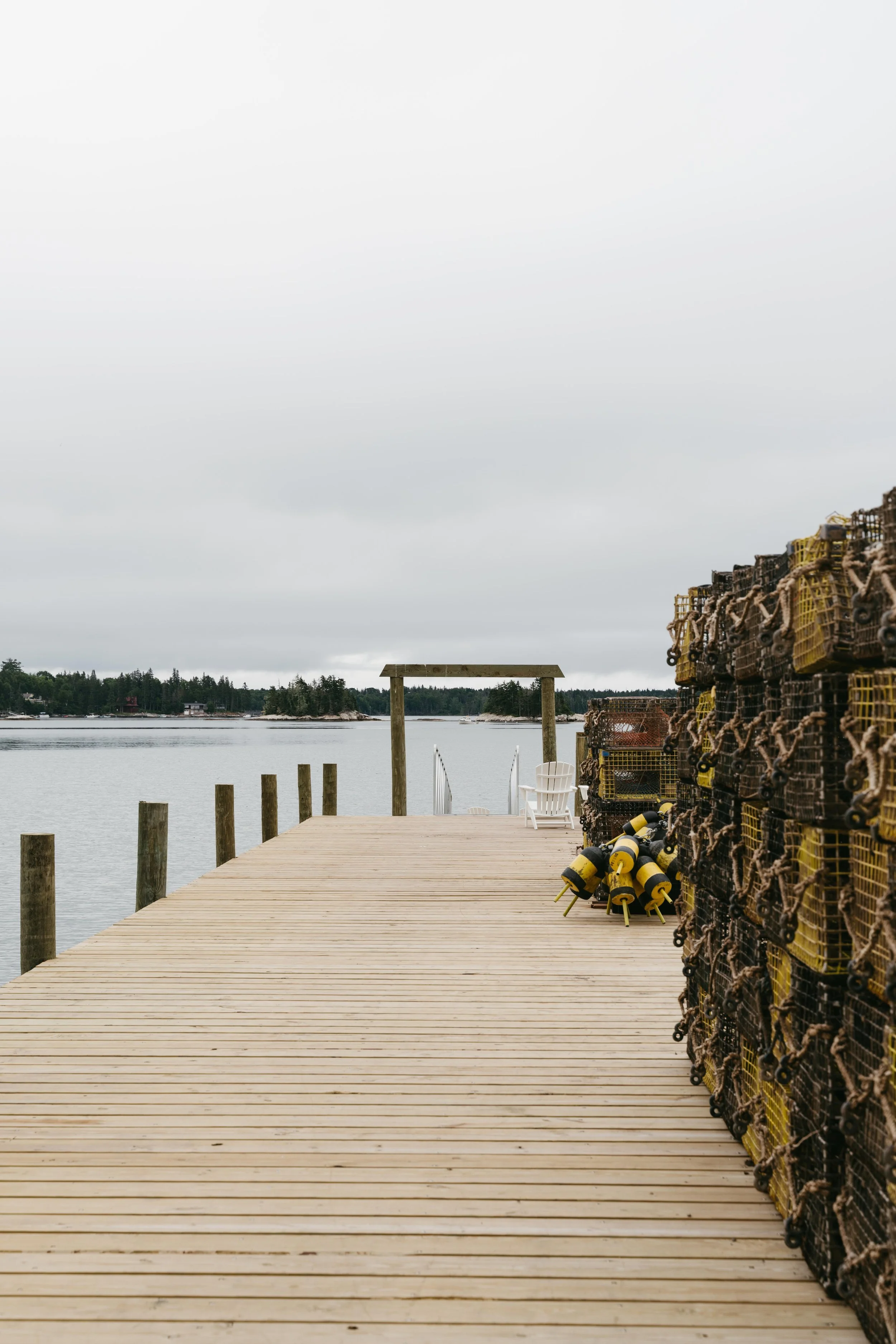 Wooden dock extending into calm water, lined with lobster traps stacked to the right, with yellow buoys, and an empty white Adirondack chair at the end, under an overcast sky.
