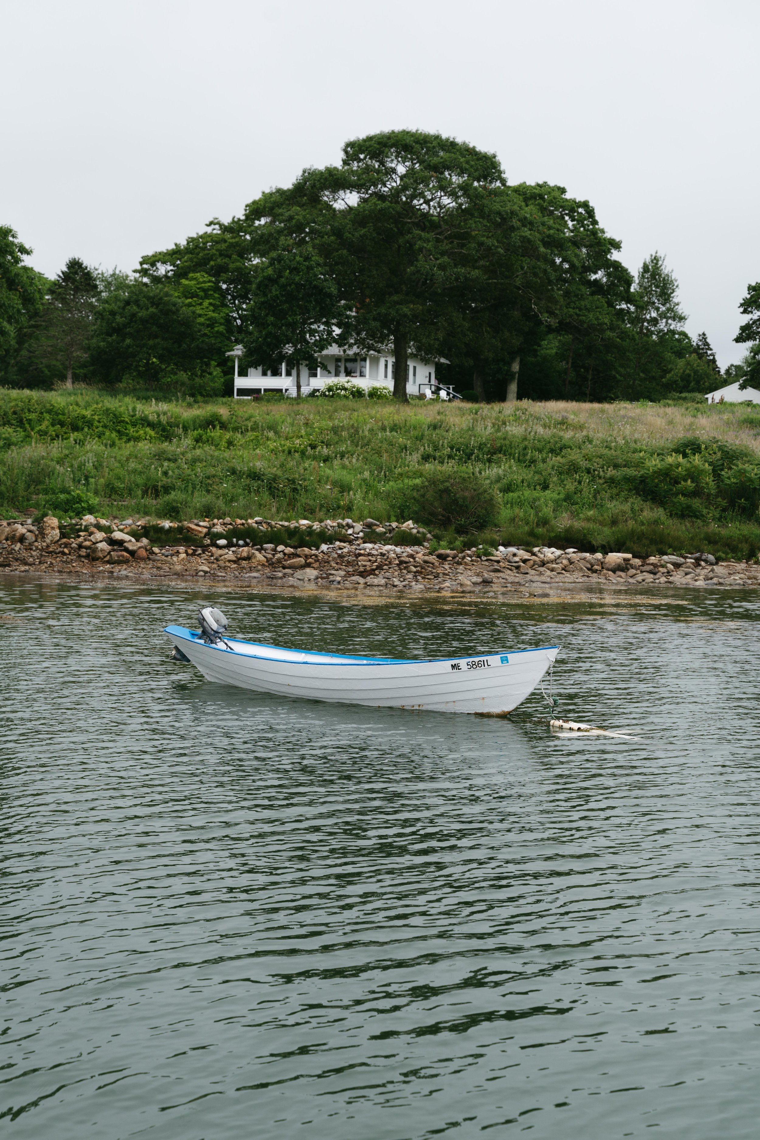 A small white boat with a blue interior floating on calm water near a rocky shoreline, with green trees and a white house in the background.