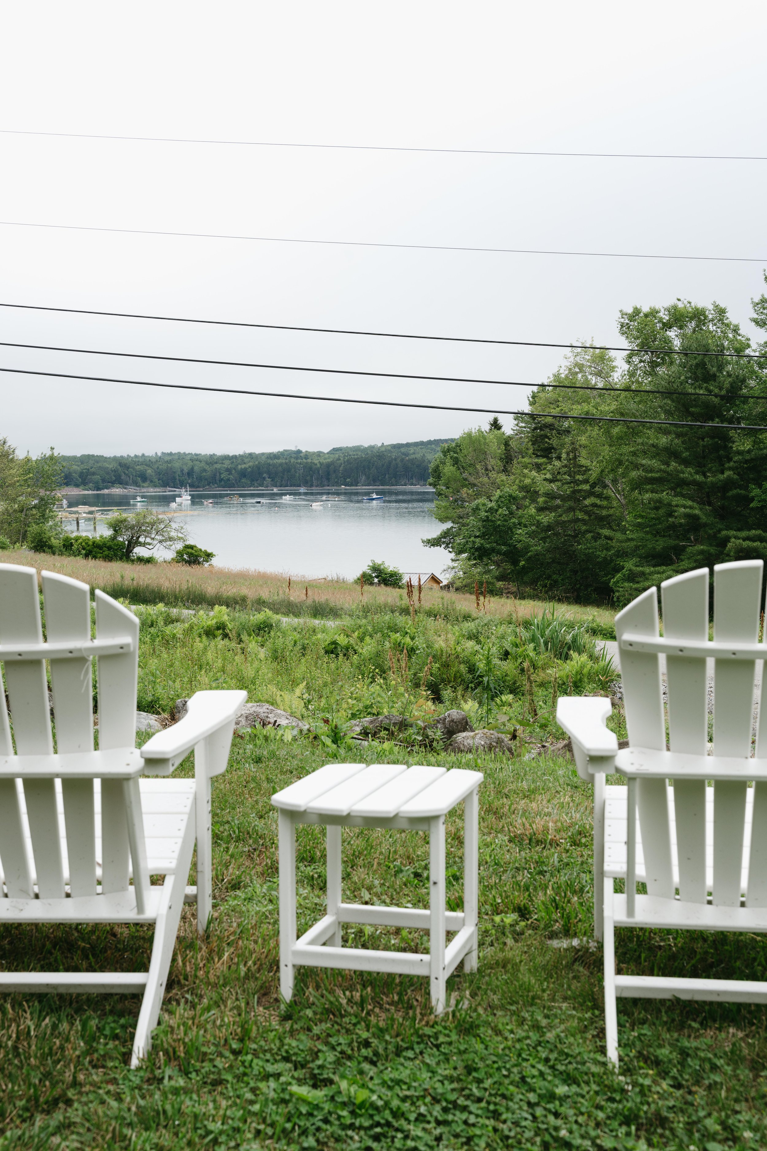 Two white Adirondack chairs with a small matching table in between on a grassy lawn overlooking a body of water with boats in the distance, surrounded by green trees and a partly cloudy sky.