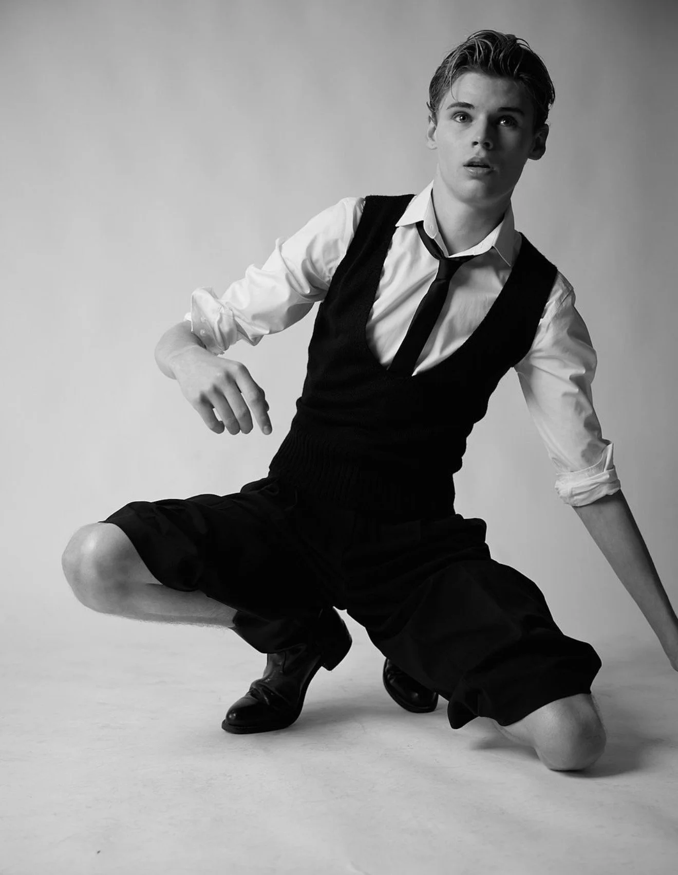 Black-and-white fashion portrait of a young man kneeling in a white shirt, black vest, tie, shorts, and dress shoes