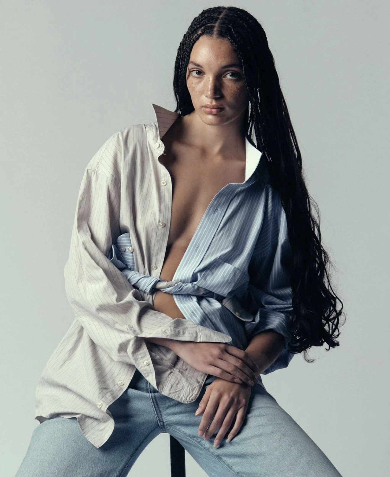 Fashion portrait of a woman with long braided hair wearing layered striped shirts and light blue jeans against a plain background