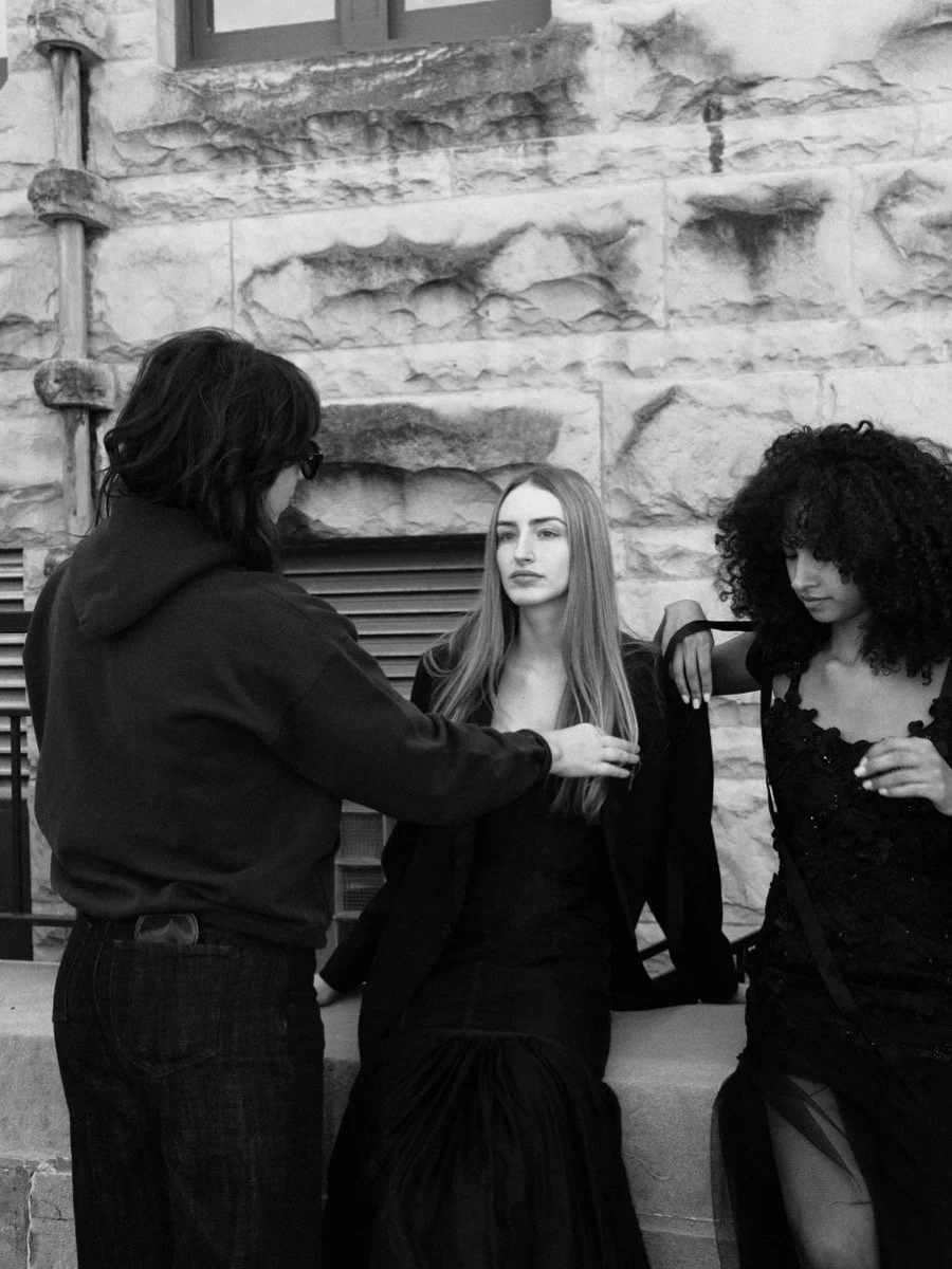 Black-and-white photo of three women in black clothing posing beside a textured stone wall, with one woman adjusting another’s hair.
