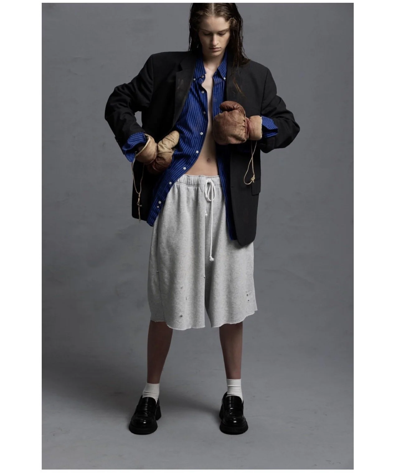Fashion model wearing a black blazer, blue striped shirt, gray sweat shorts, boxing gloves, white socks, and black loafers