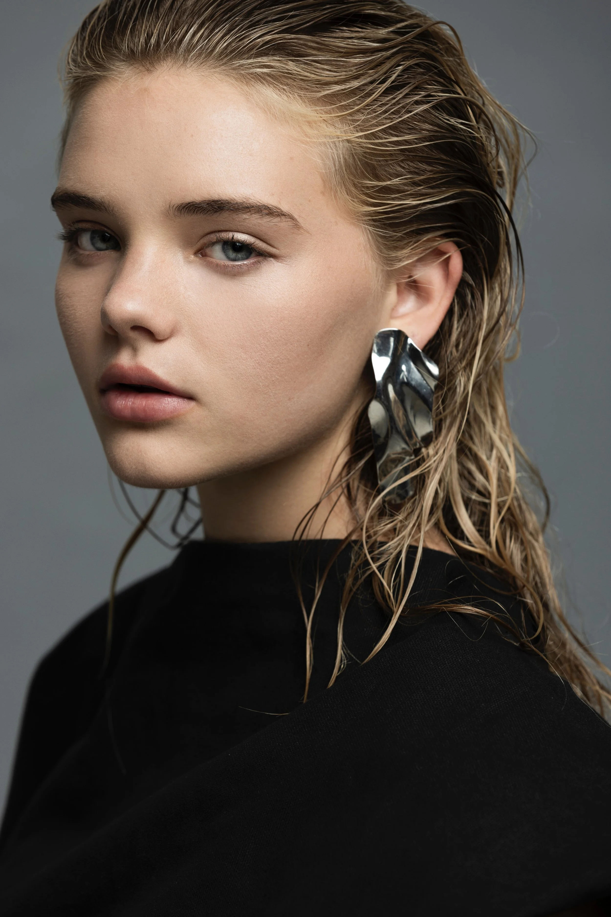 Close-up portrait of a blonde woman with blue eyes, wet-look hair, and a large metallic earring against a gray background.