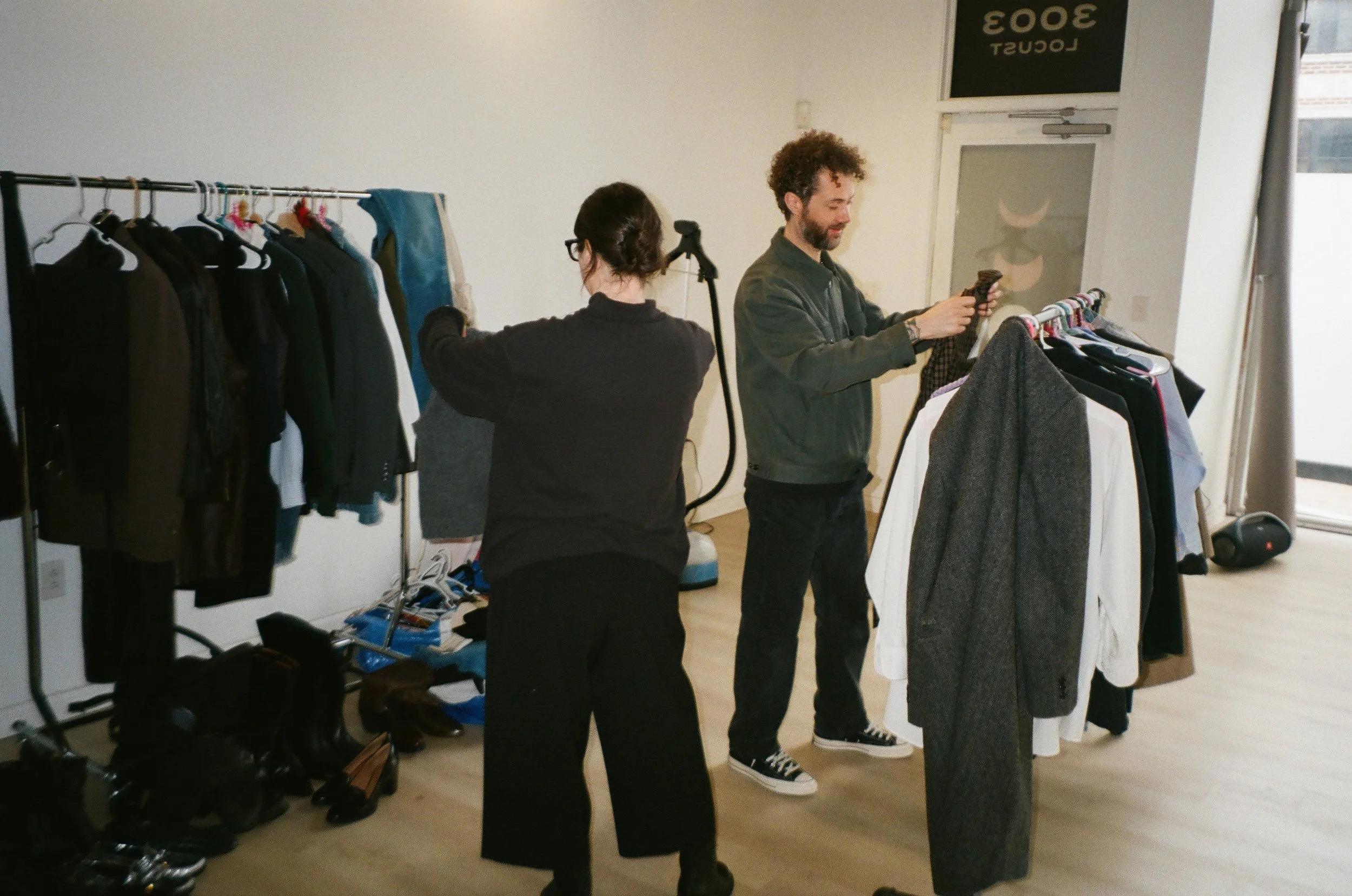 Two people shopping for clothes in a boutique, with racks of clothing and shoes around them.