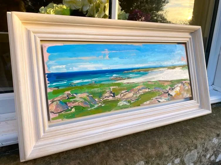 An abstract landscape painting of a beach with greenery and rocks in the foreground, blue sky, and ocean waves in the background, framed in a white wooden frame, leaning against a window.