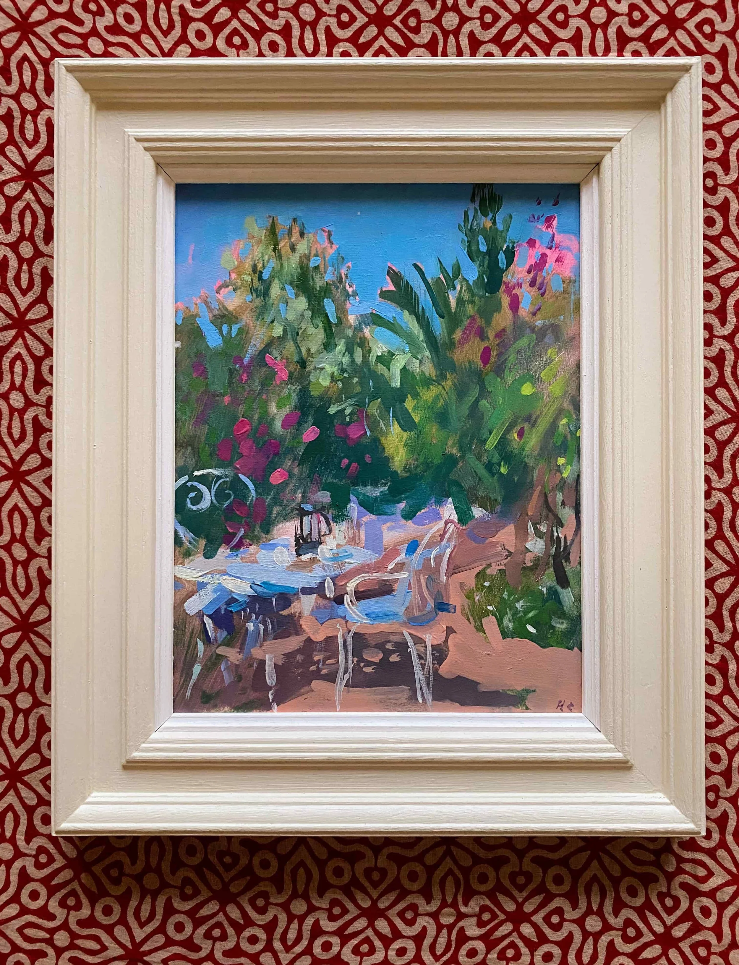 A colorful landscape painting of lush green bushes and trees with a bright blue sky background, framed in a white wooden frame, and placed against a red patterned wall.