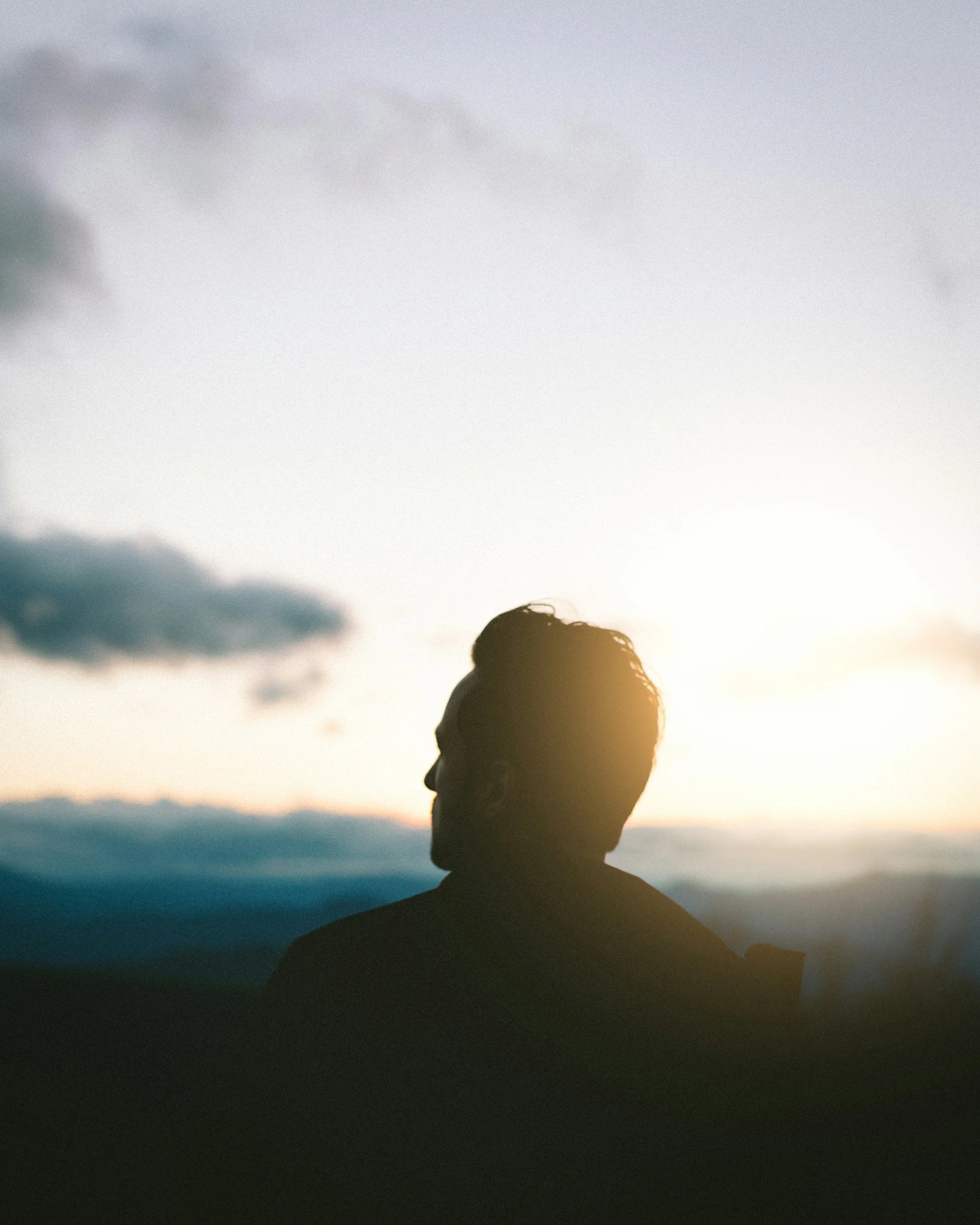 Silhouette of a man with short hair against a sunset sky, overlooking distant mountains.