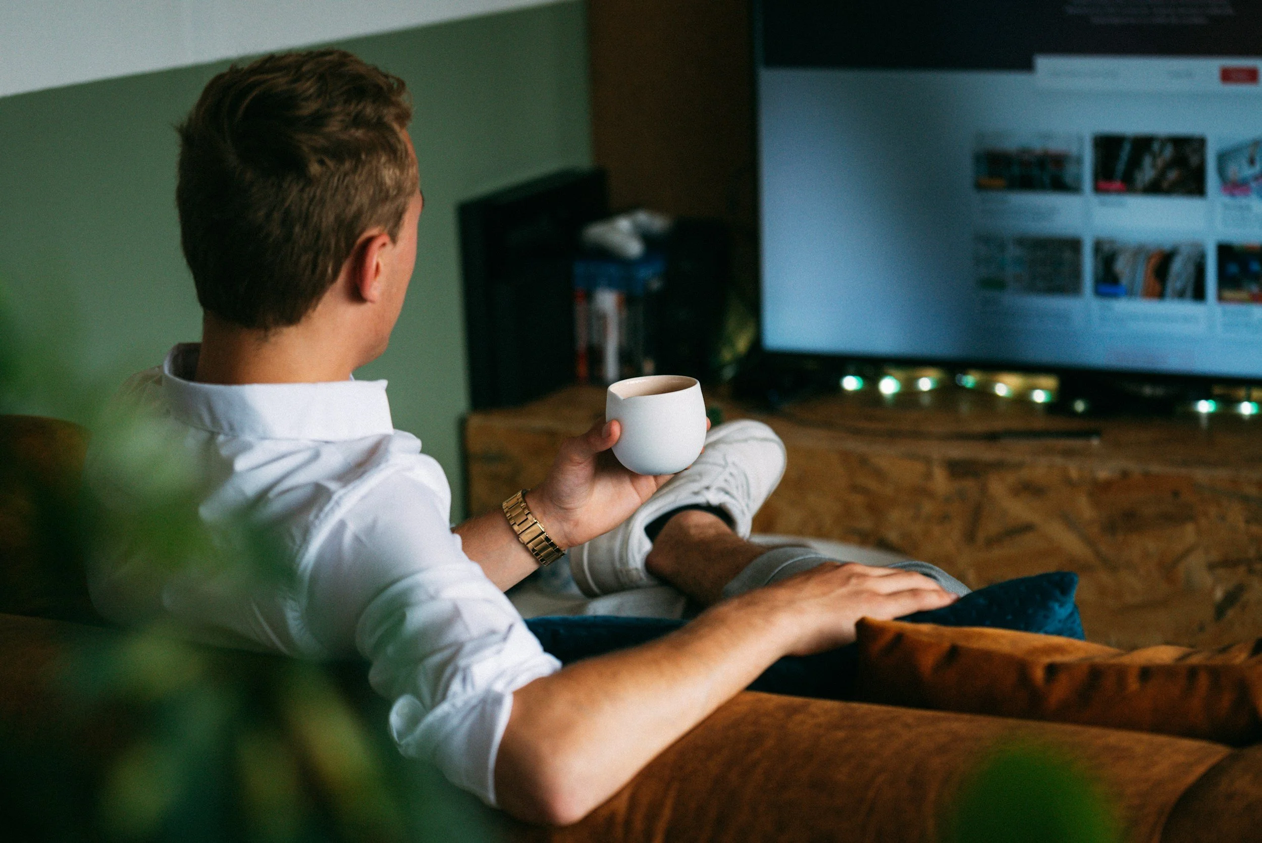 A man with light skin and brown hair, wearing a white shirt and a gold watch, sitting on a sofa with a coffee mug, watching a television in a living room.