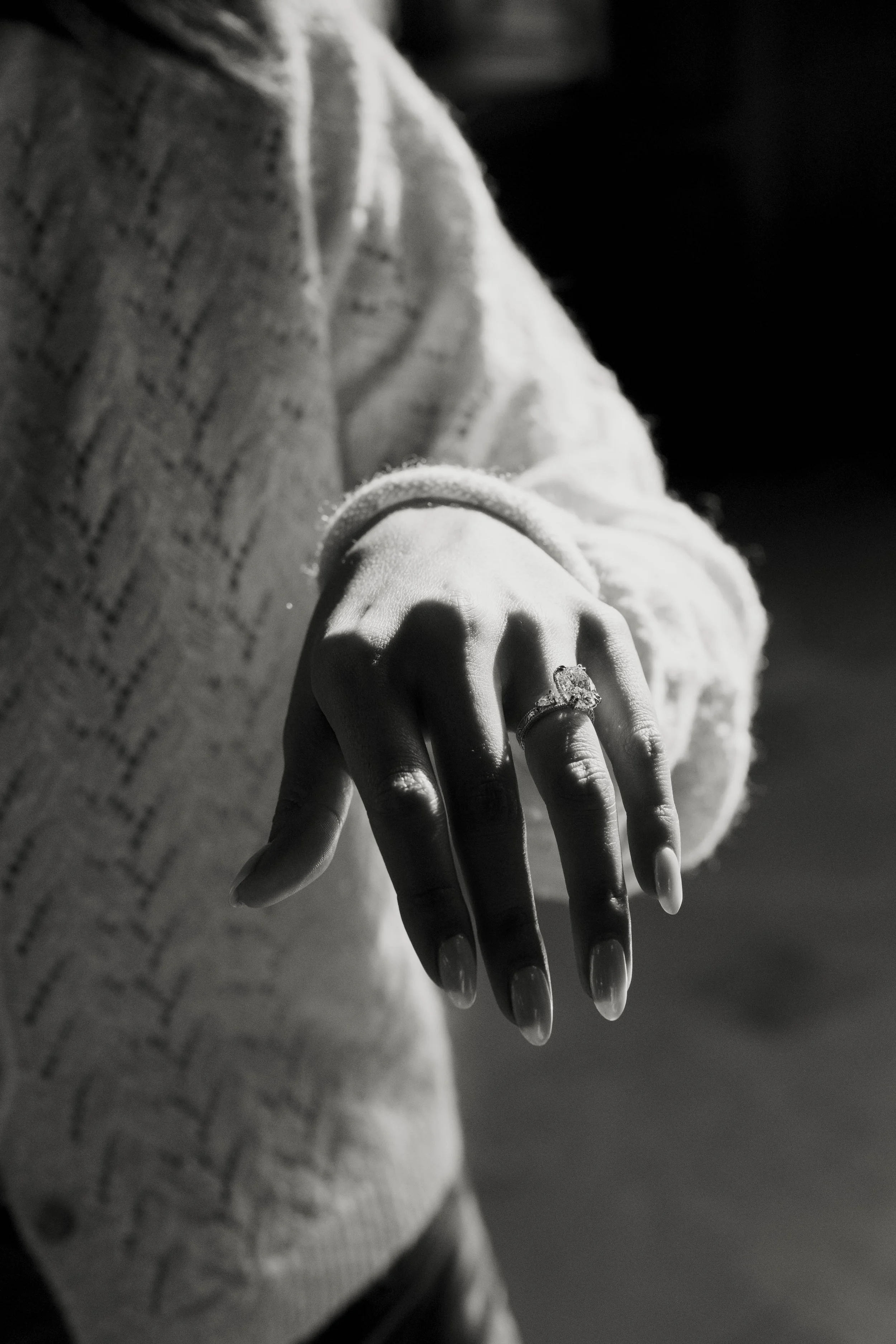 A person wearing a sweater shows their left hand with an engagement ring on the ring finger, in black and white.