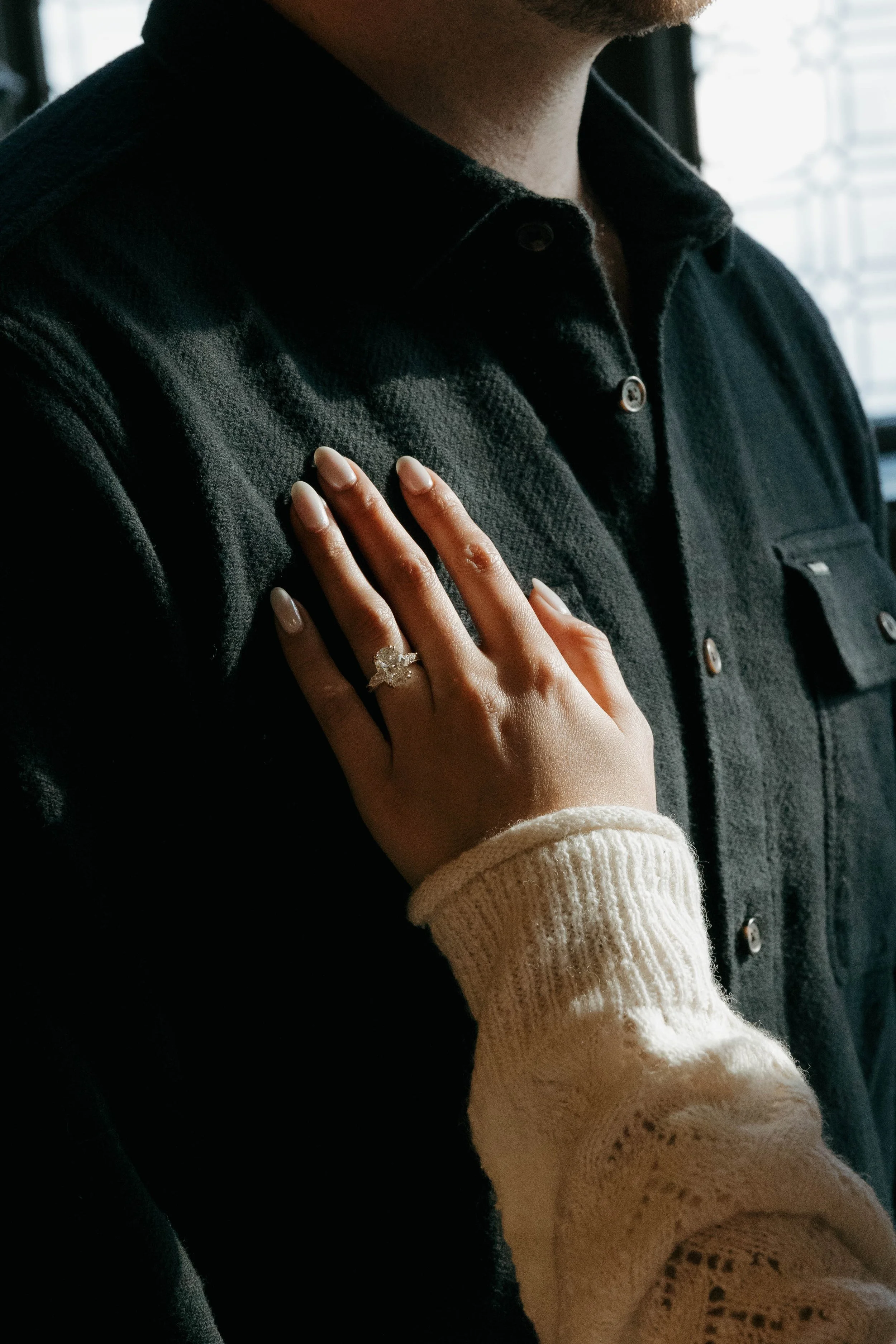 A woman placing her hand on a man's chest, showing an engagement ring with a large gemstone. The man is wearing a dark button-up shirt, and the woman is wearing a cream-colored, knitted sweater.