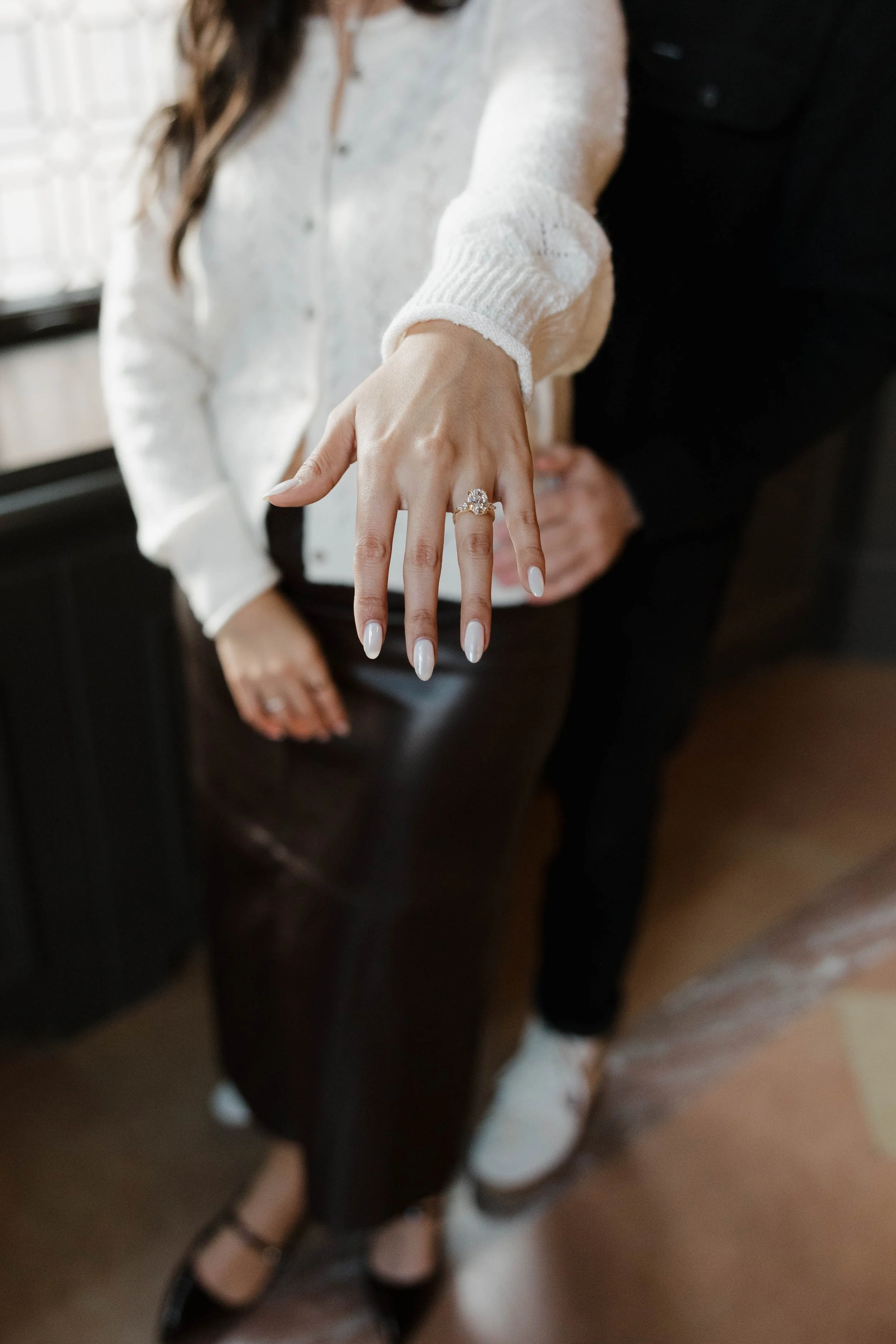A woman displays her wedding ring on her left hand, with a man standing behind her. The woman is wearing a white sweater, black pants, and black heels, while the man is dressed in dark clothing. The focus is on the woman's hand and ring.