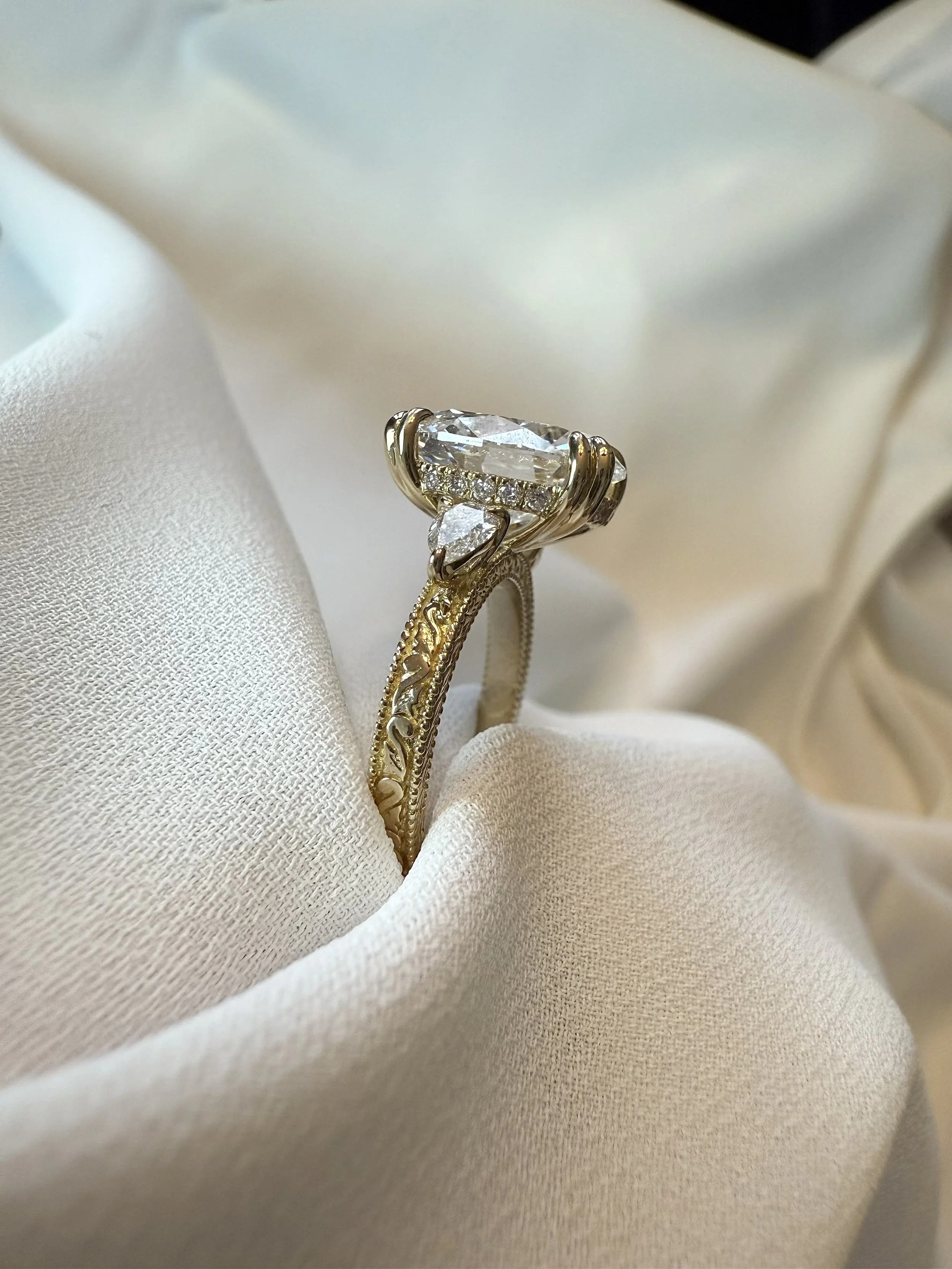 Close-up of a gold engagement ring with a large central diamond and smaller diamonds, resting on white satin fabric.