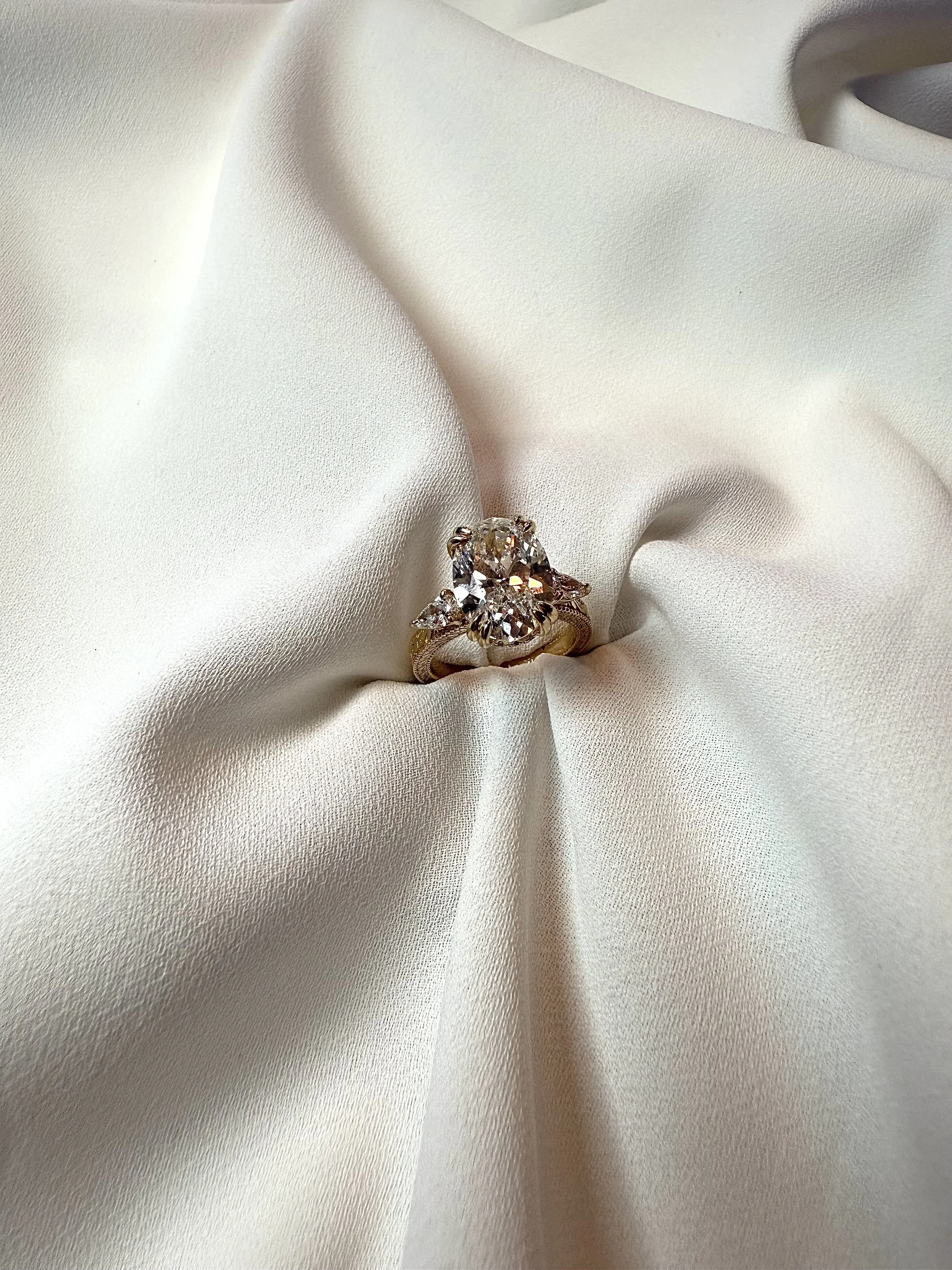 A diamond solitaire engagement ring with a central round cut diamond surrounded by smaller diamonds, set on a gold band, placed on white satin fabric.