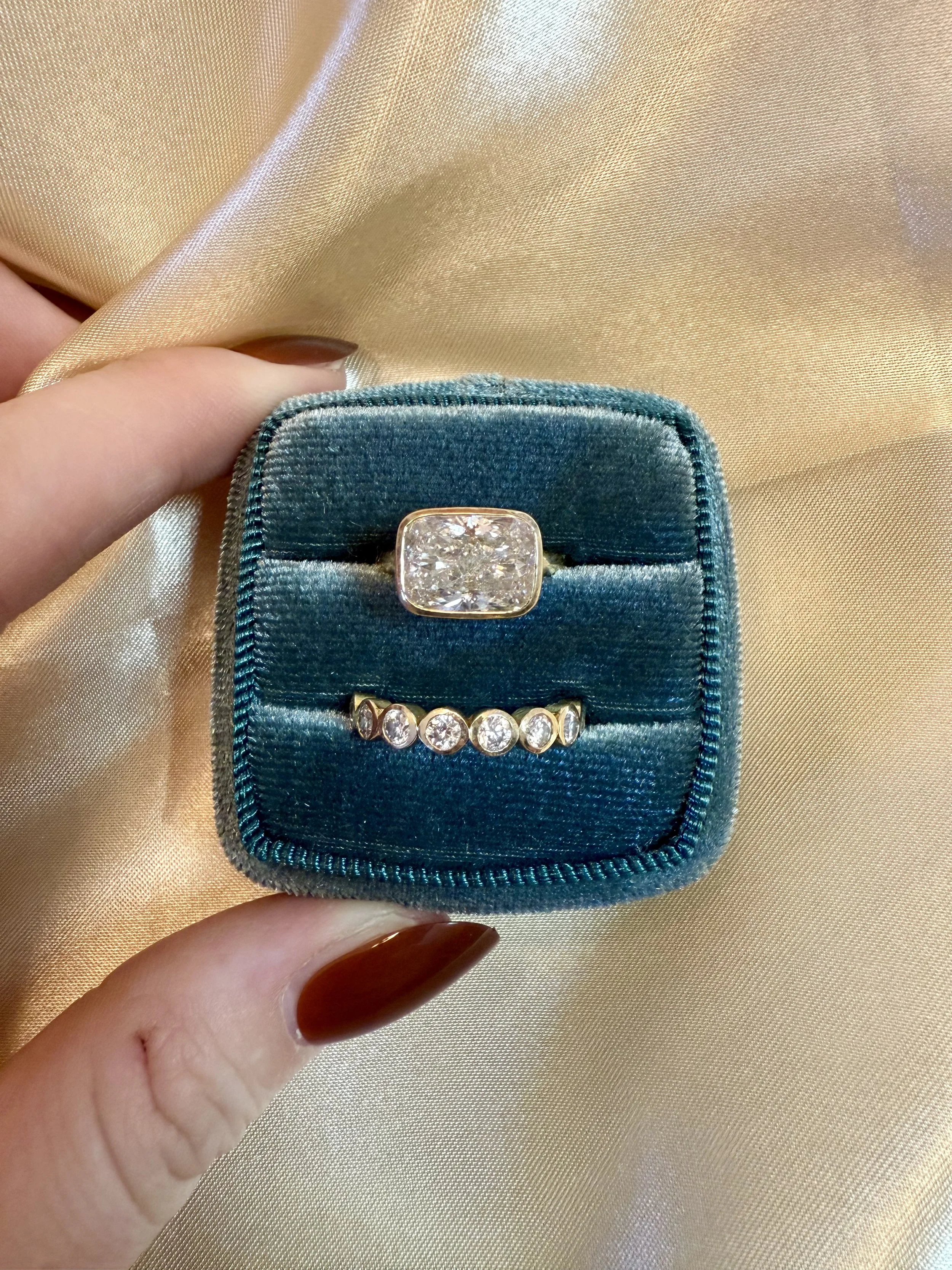 A ring with a large square-shaped diamond in a velvet jewelry box, with a smaller gold ring with multiple small round diamonds, against a gold satin background.