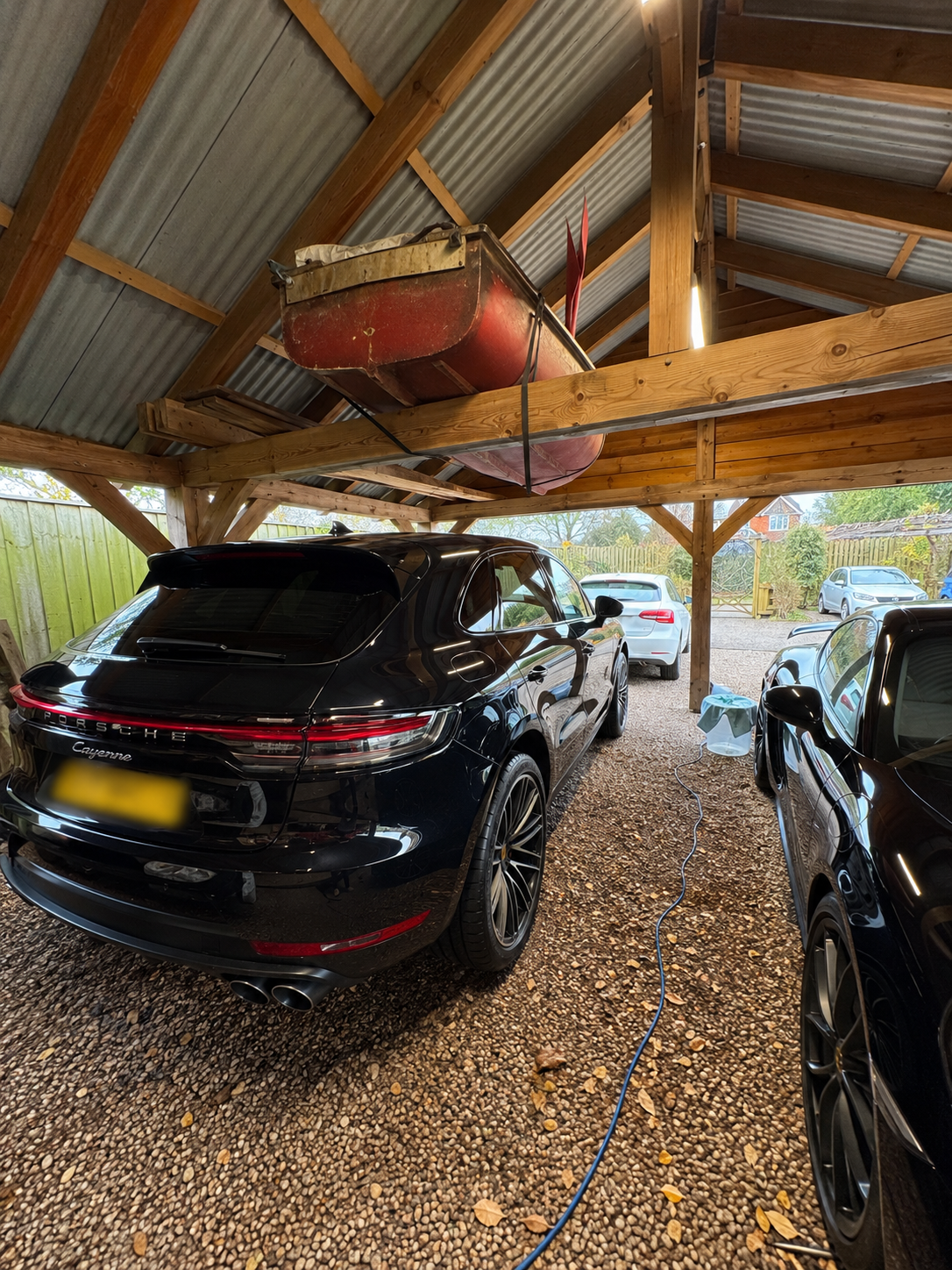 A black Porsche Cayenne parked under a wooden and metal roof, with an old red boat stored above it, inside a carport with gravel floor and other cars parked outside.
