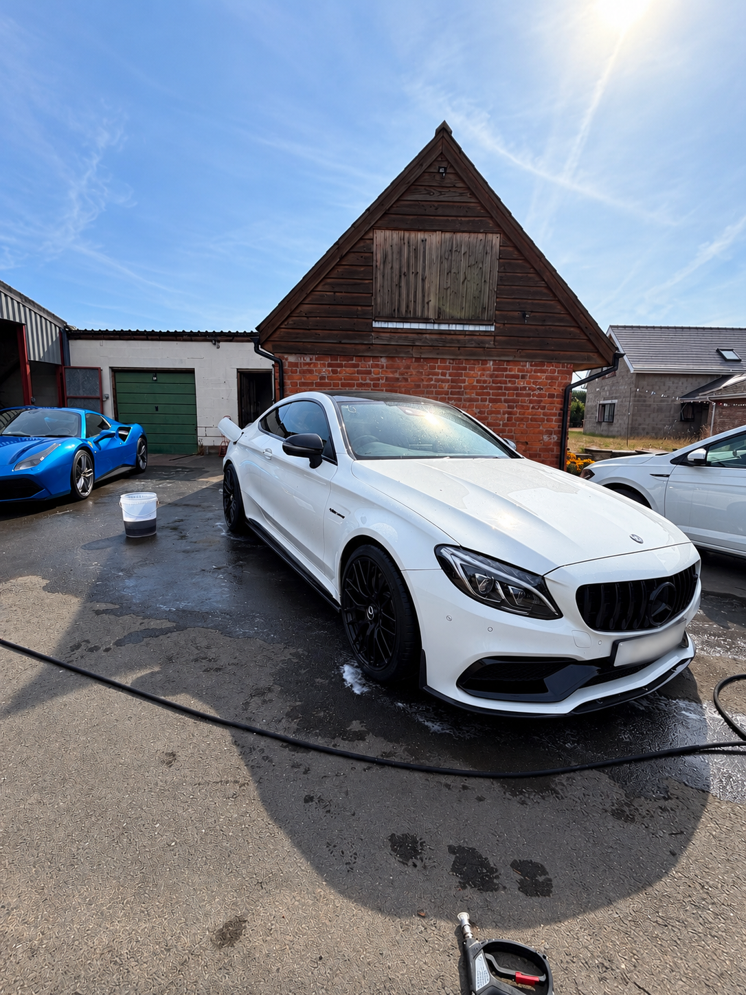 A white Mercedes-Benz car is being washed in a driveway with soap suds and a bucket, with a blue sports car and other vehicles in the background under a blue sky.