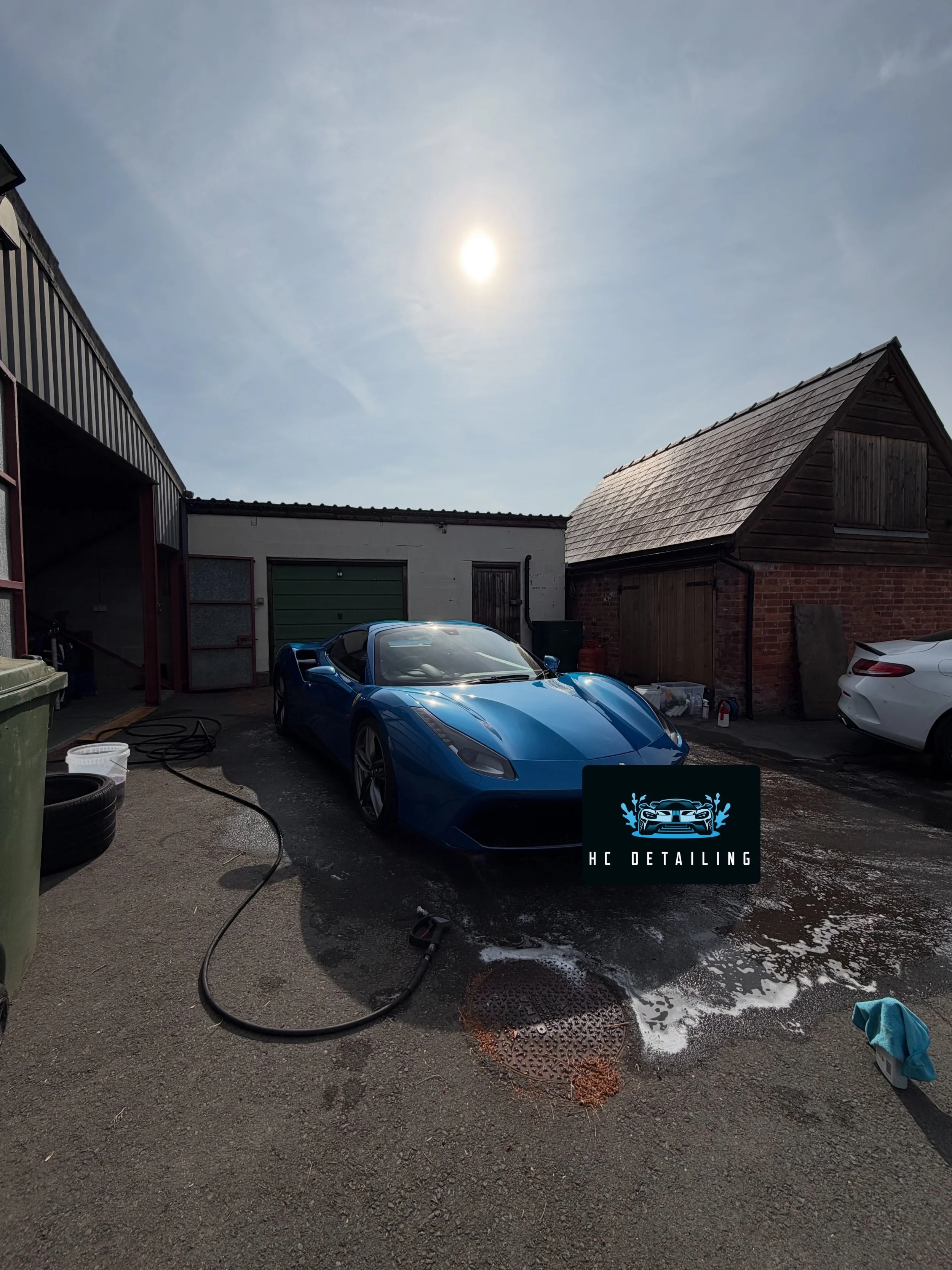 A blue sports car being washed outdoors on a driveway, with soap suds on the ground, surrounding garage buildings, and a bright sun in the clear sky.