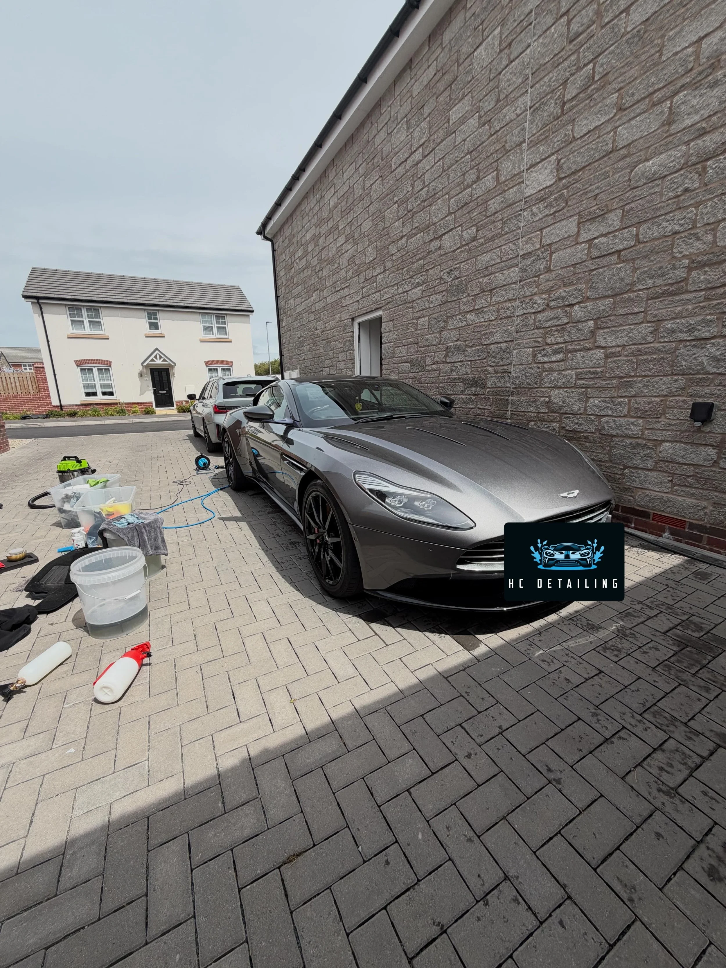 A gray sports car parked on a paved driveway in front of a brick house with work tools and supplies scattered around.